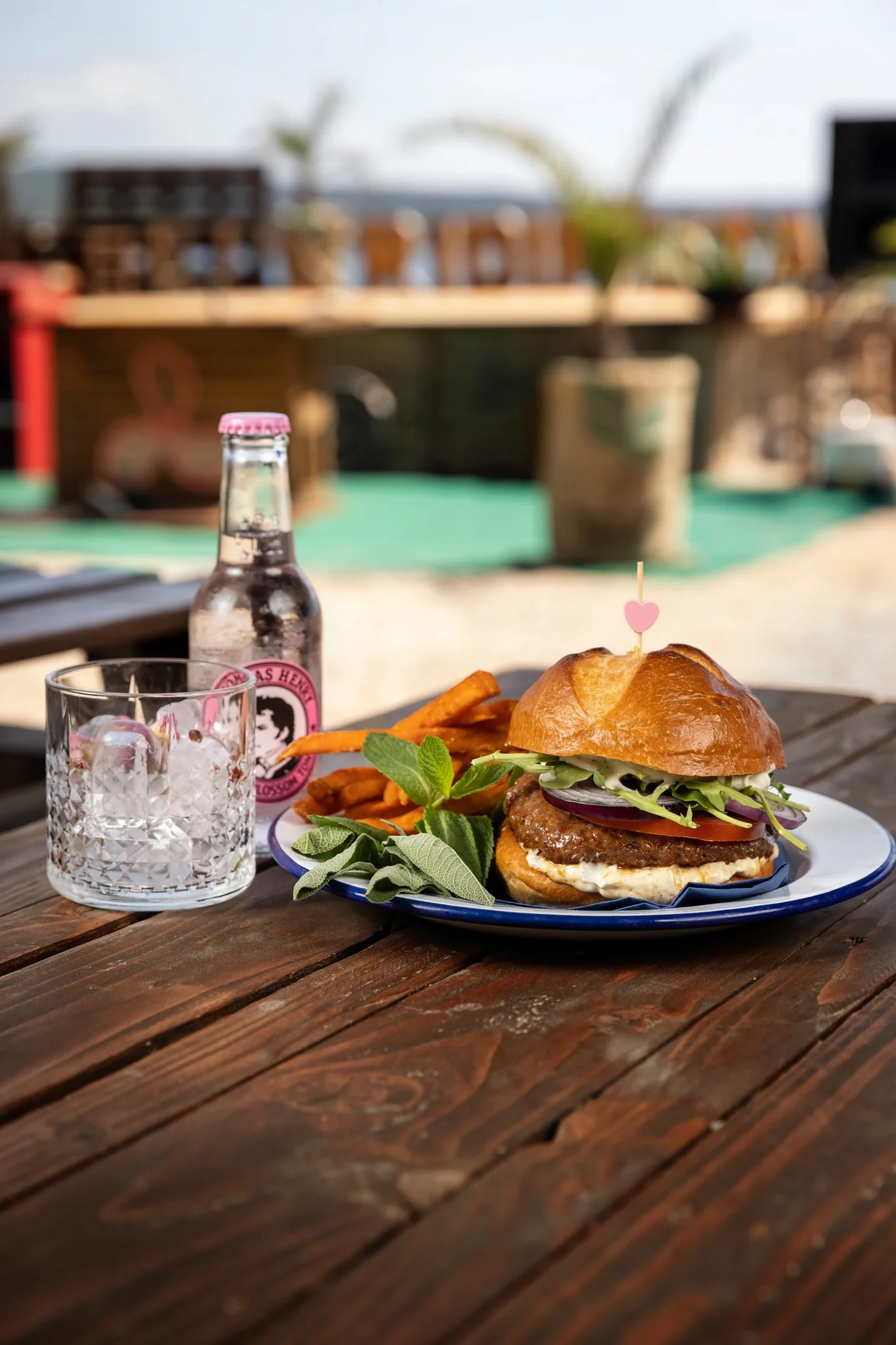 A hamburger with lettuce, tomato, onion, and sauce on a bun, served on a blue plate with fresh herbs. A glass of ice water, a bottled beverage with a pink cap, and a side of sweet potato fries are on a rustic wooden table. In the background, outdoor 