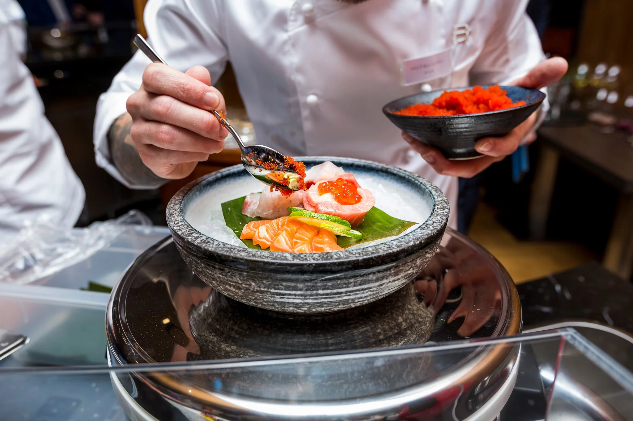 A chef in a white uniform adds roe to a seafood dish with sashimi and garnishes on a stone plate.