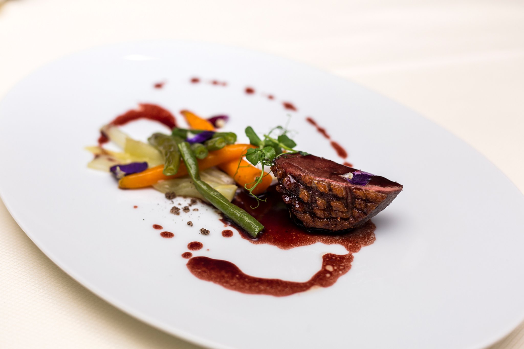 A gourmet dish featuring a piece of glazed, sliced meat with a rich red sauce on a white plate, accompanied by a colorful assortment of vegetables including carrots, green beans, and garnished with edible flowers.