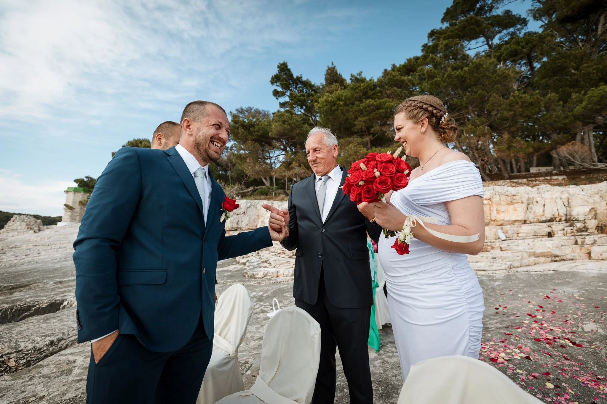 A wedding ceremony taking place outdoors by the water, with a groom in a blue suit and a bride in a white dress holding a bouquet of red roses, smiling at each other with a officiant standing between them.