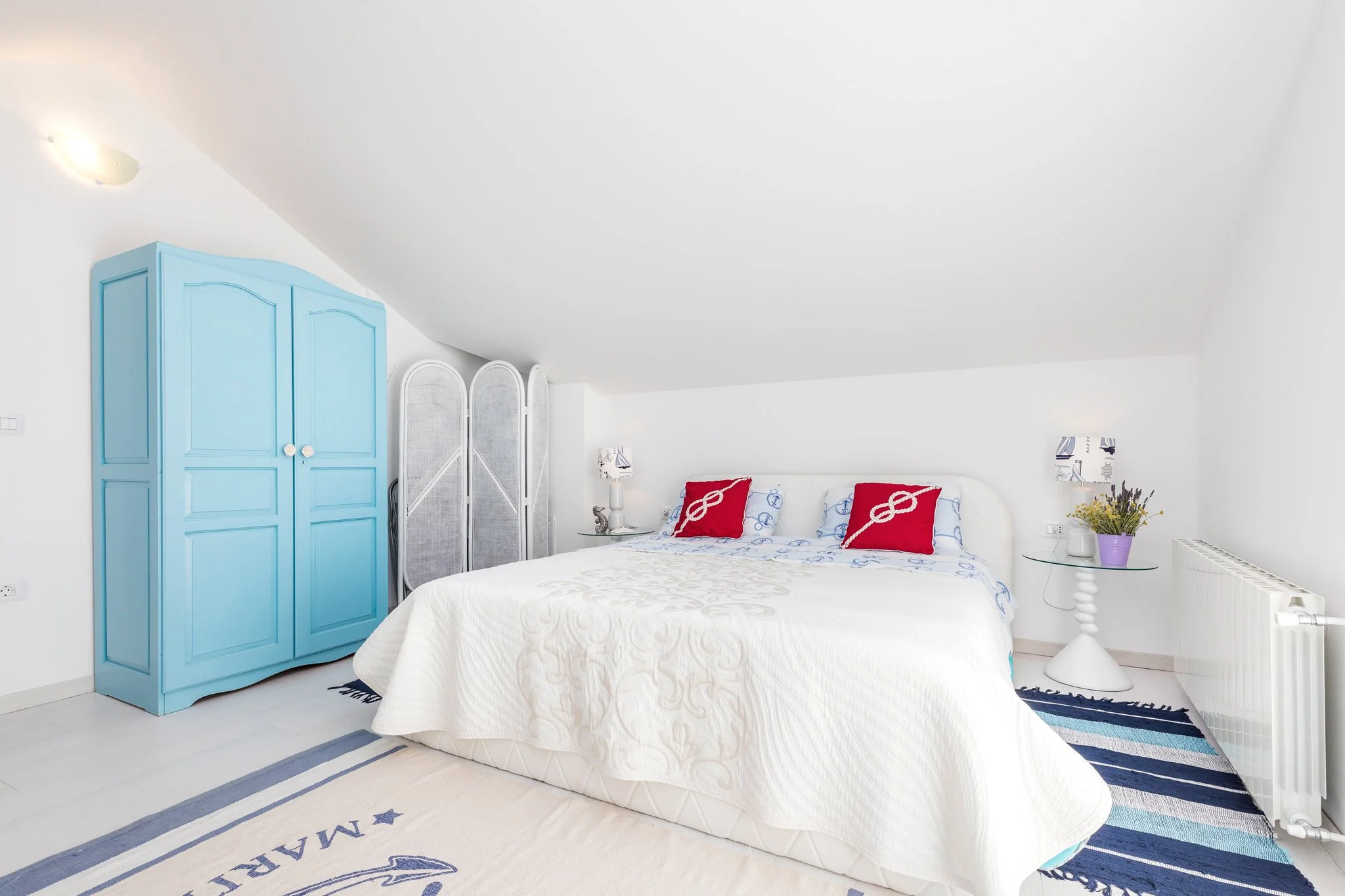 A bedroom with a white bed, blue wardrobe, and decorative pillows with nautical motifs, a side table with a vase of flowers, and wall lamps.