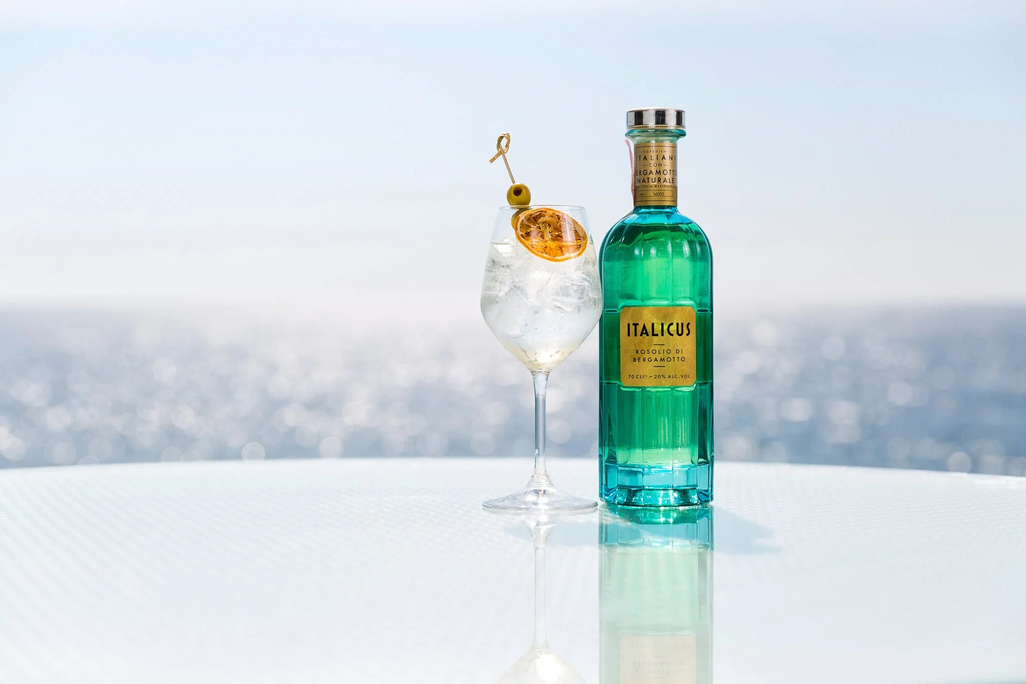 A bottle of Italicus Rosolio di Bergamotto liqueur next to a glass filled with ice, a slice of orange, and an olive on a pick, set against a bright background with a blurred seascape.