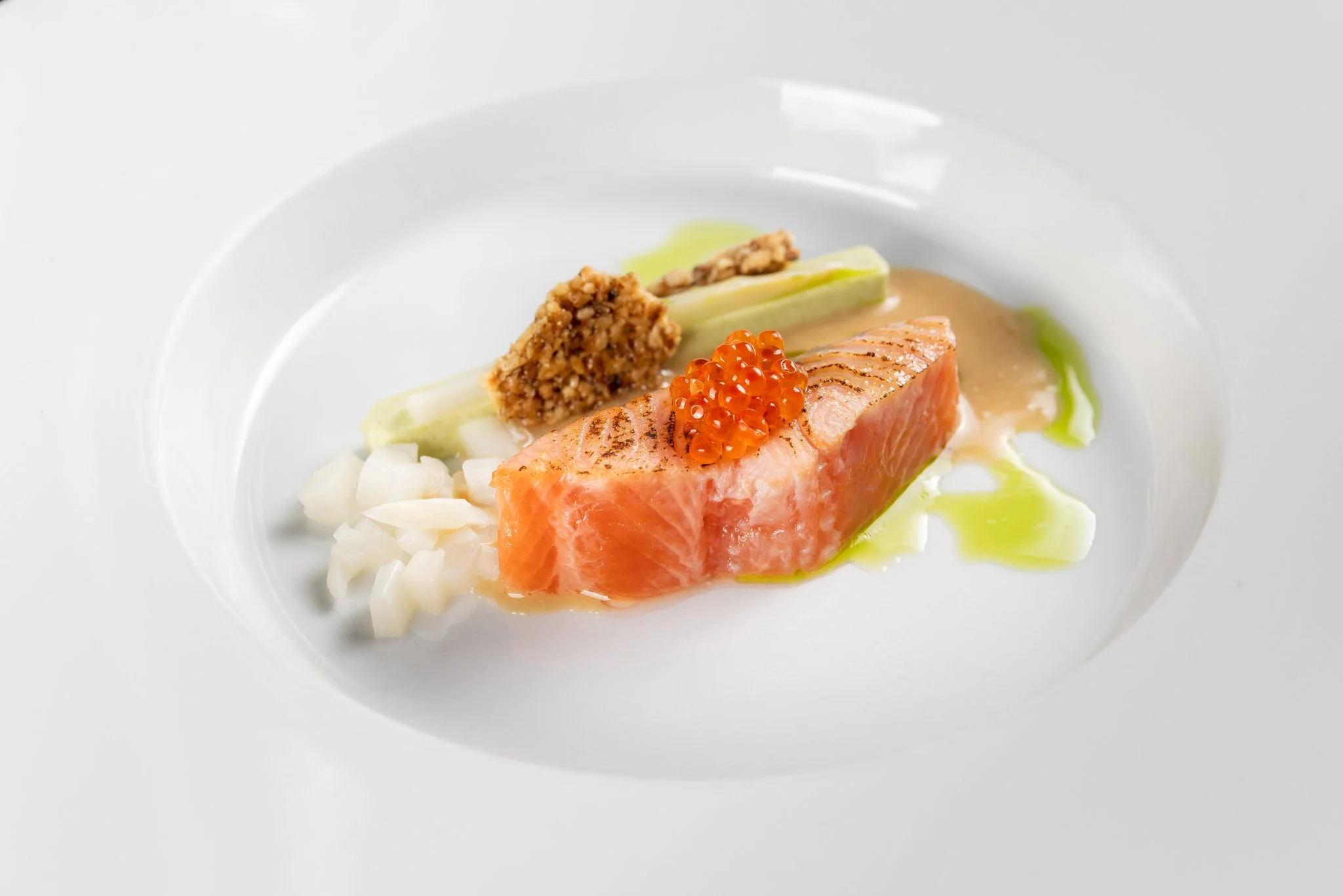A plated dish featuring seared salmon, topped with orange fish roe, accompanied by diced onions, a piece of celery, a crispy breadcrumb or cracker, and a sauce or broth on a white plate.