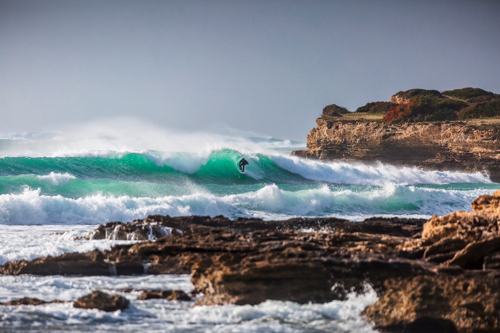 A person surfing a large green wave near rocky shore with a hillside covered in vegetation in the background.