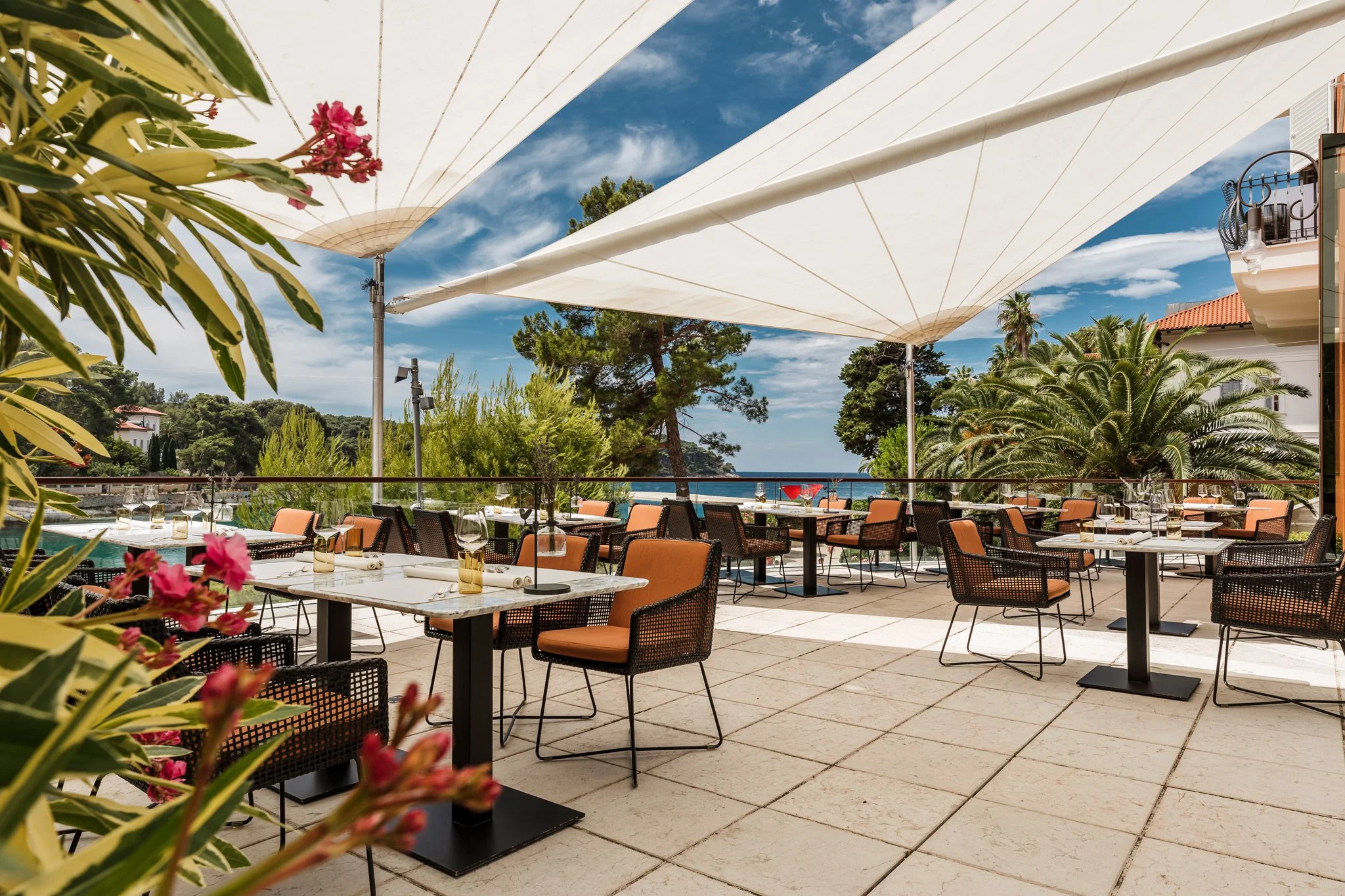 An outdoor restaurant patio with tables and chairs, large white canopy shades, lush green trees, and a view of the sky and ocean in the background.Alfred Keller in Mali Lošinj, Boutique Hotel Alhambra.