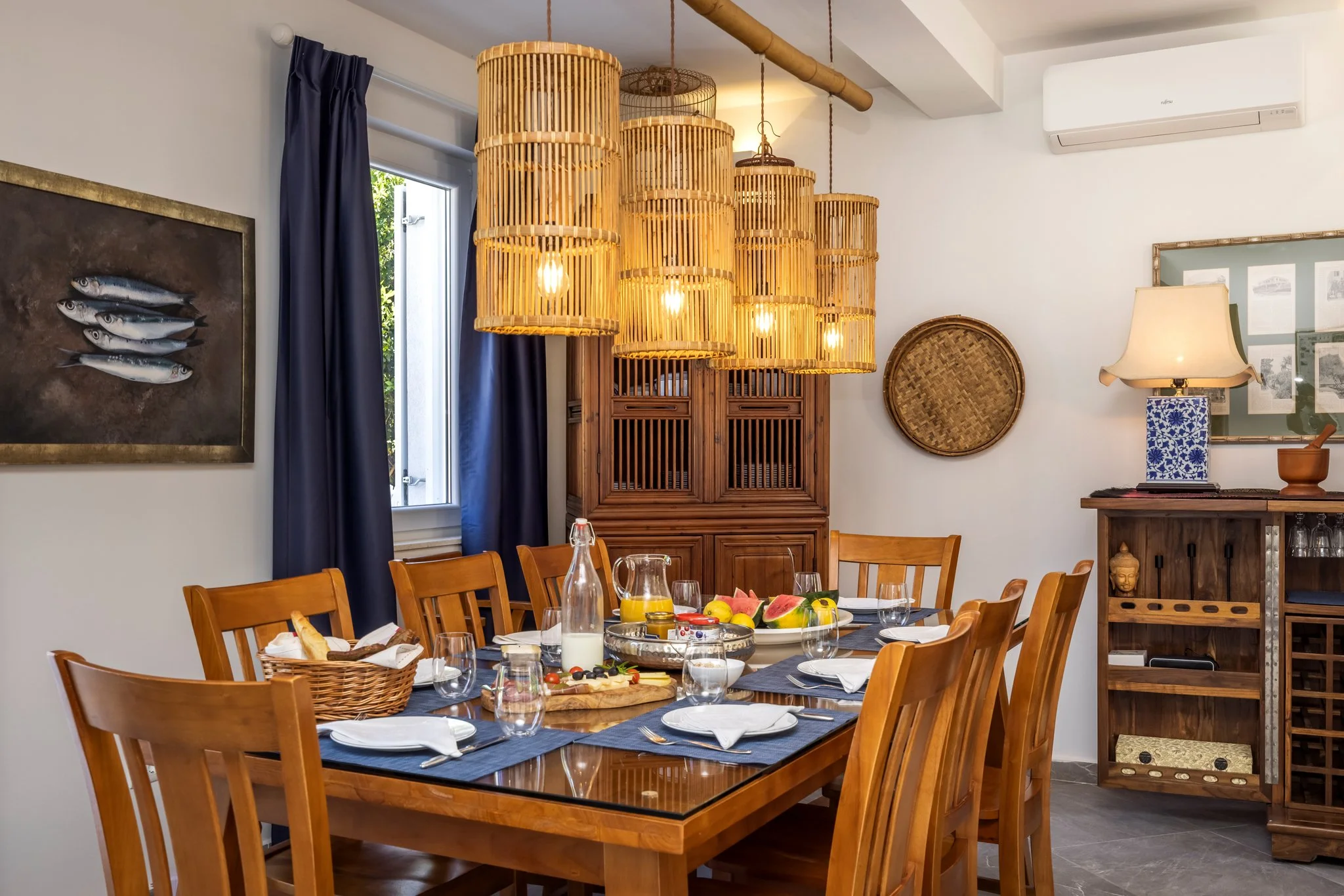 A dining room with wooden chairs around a table set with plates, glasses, and a fruit platter. Hanging bamboo pendant lights above the table. A wall with a painting of fish, a window with blue curtains, and various decorative items including a wicker