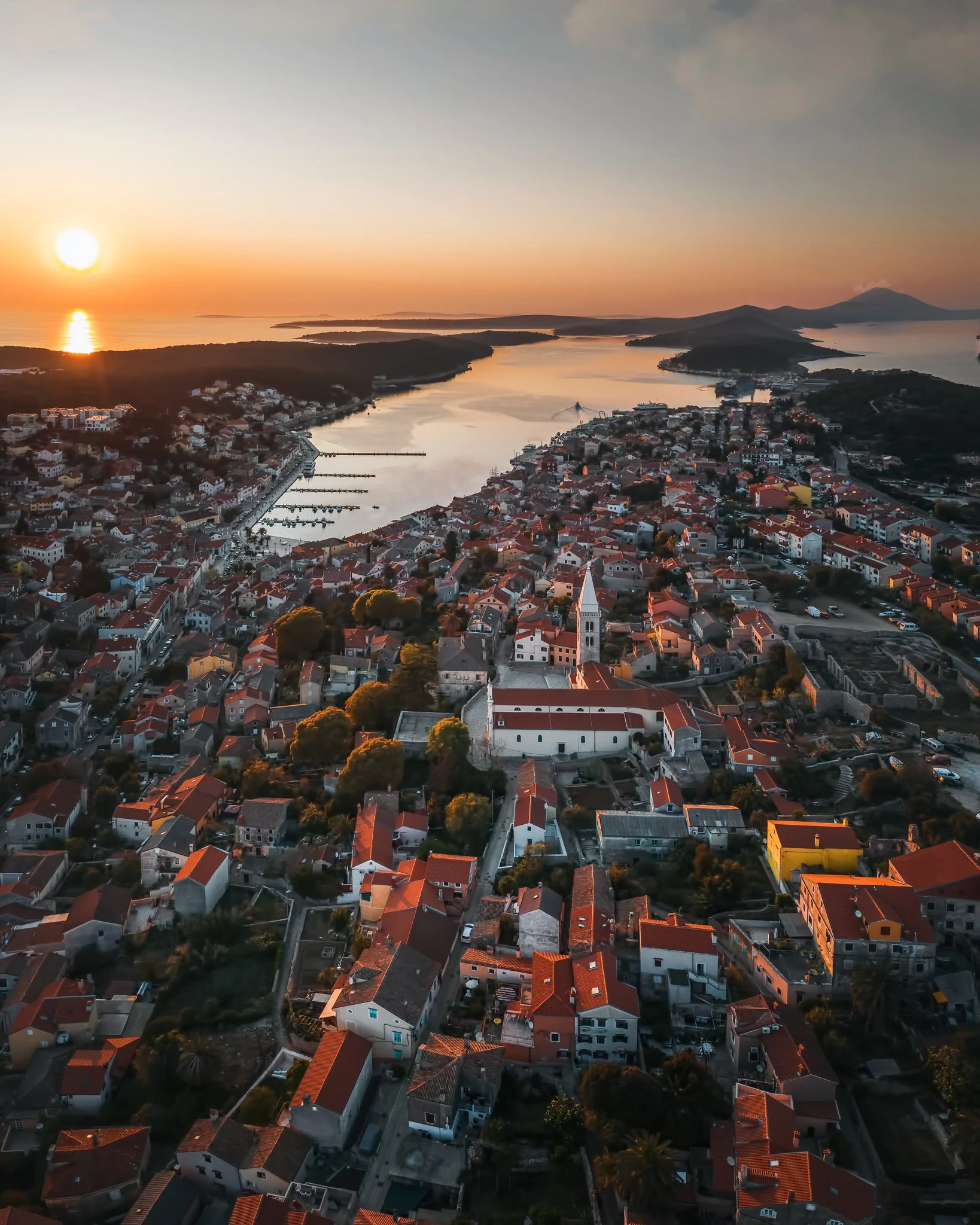 An aerial view of a coastal town during sunset, showing buildings with red-tiled roofs, a winding waterway, surrounding hills, and a mountain in the distance.