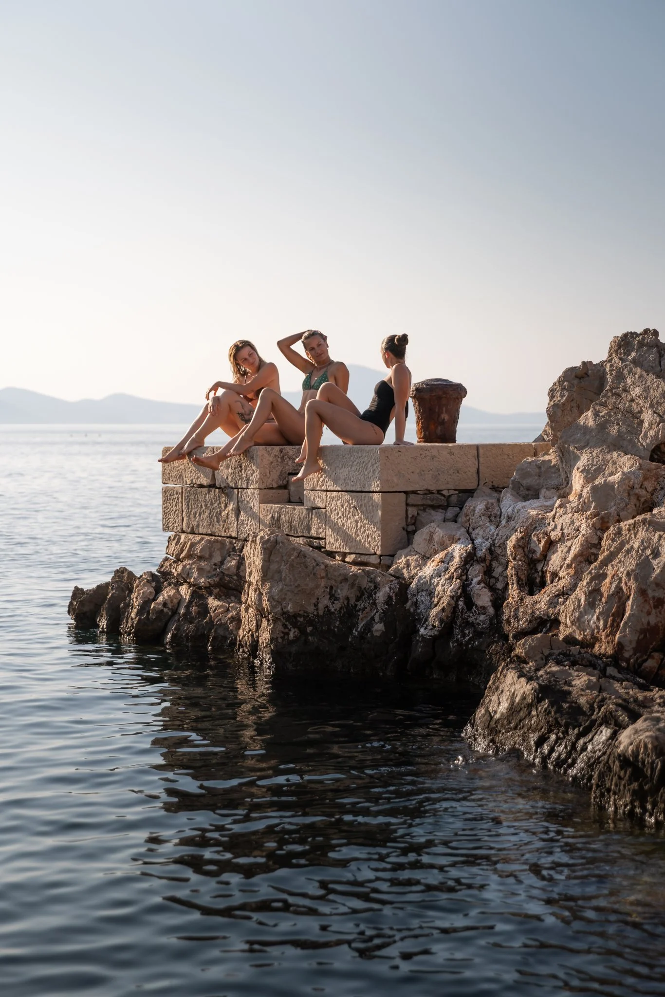 Three women sitting on rocks beside the water, with mountain and sky in the background, during sunset or sunrise.