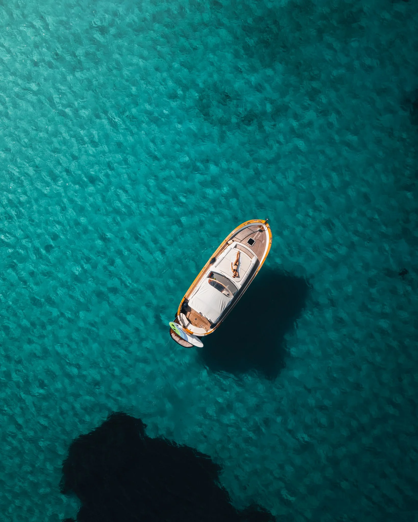 A boat floating on clear, blue-green ocean water with rocky shoreline shadows visible at the bottom of the image.