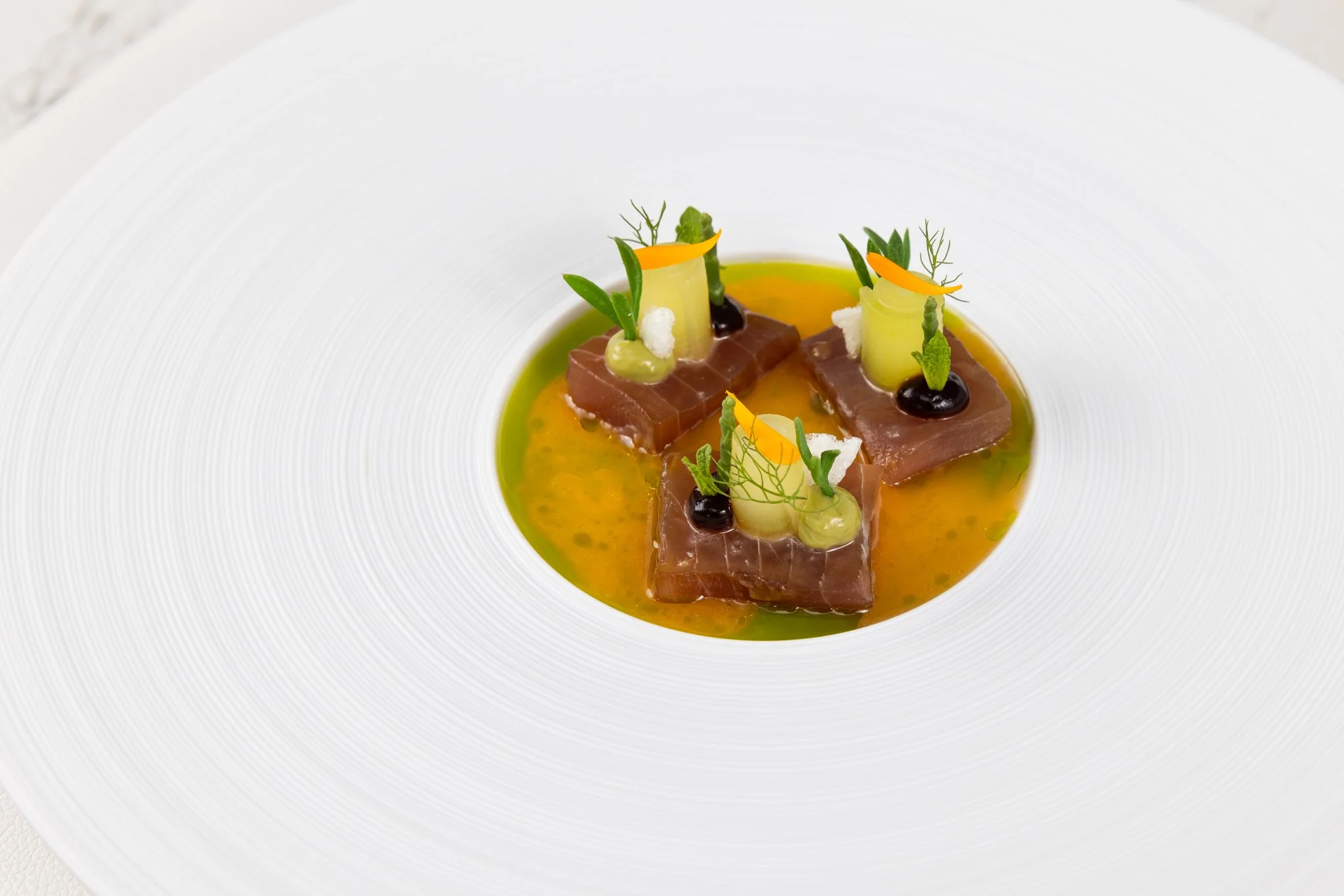 Elegant plated dish featuring slices of seared tuna topped with various garnishes, including asparagus, microgreens, and small dollops of sauces, served in a vibrant yellow sauce on a white textured plate.