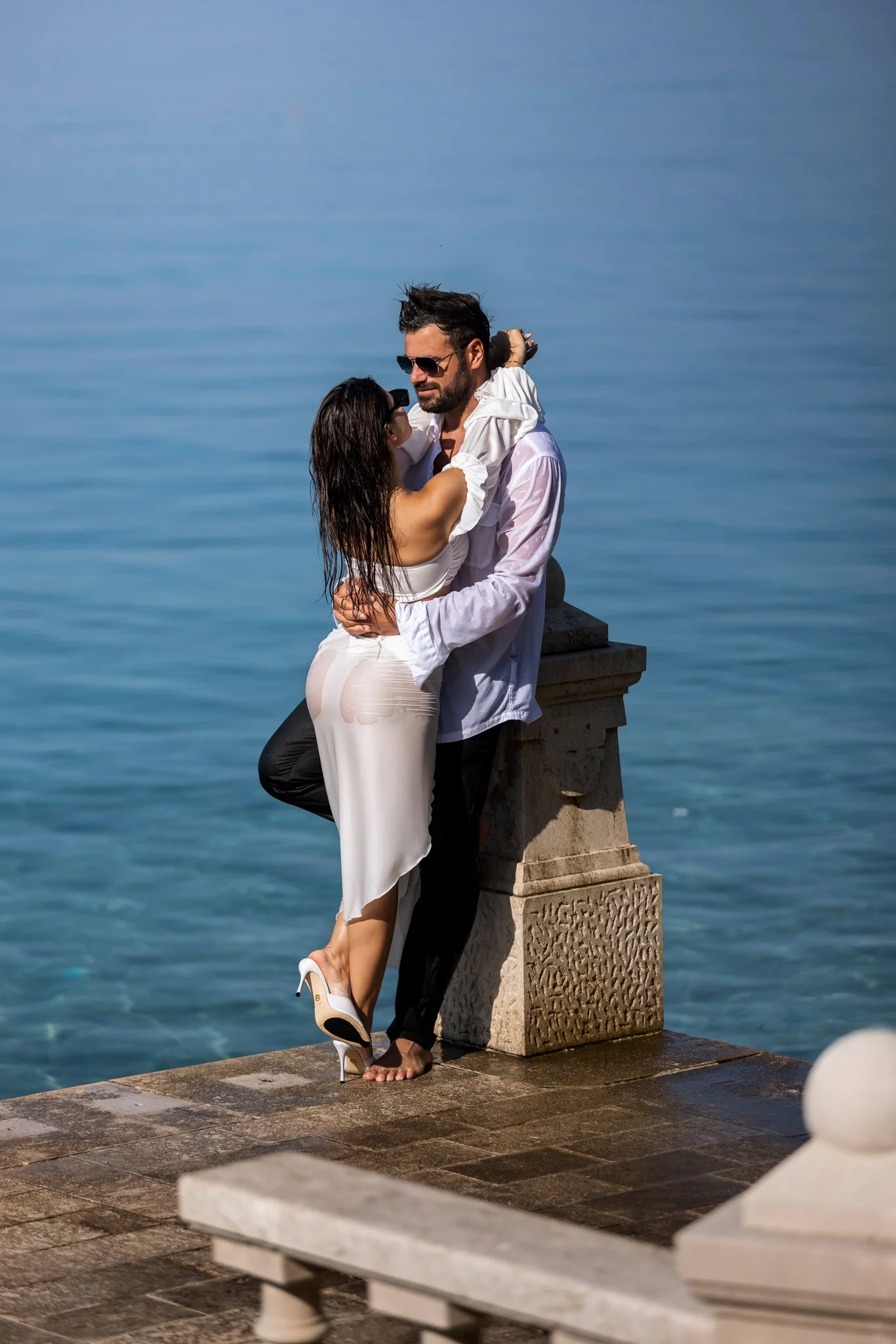 A man and woman embrace by the water on a stone pier, with the woman wearing high heels and a white dress, and the man barefoot, both wearing sunglasses.