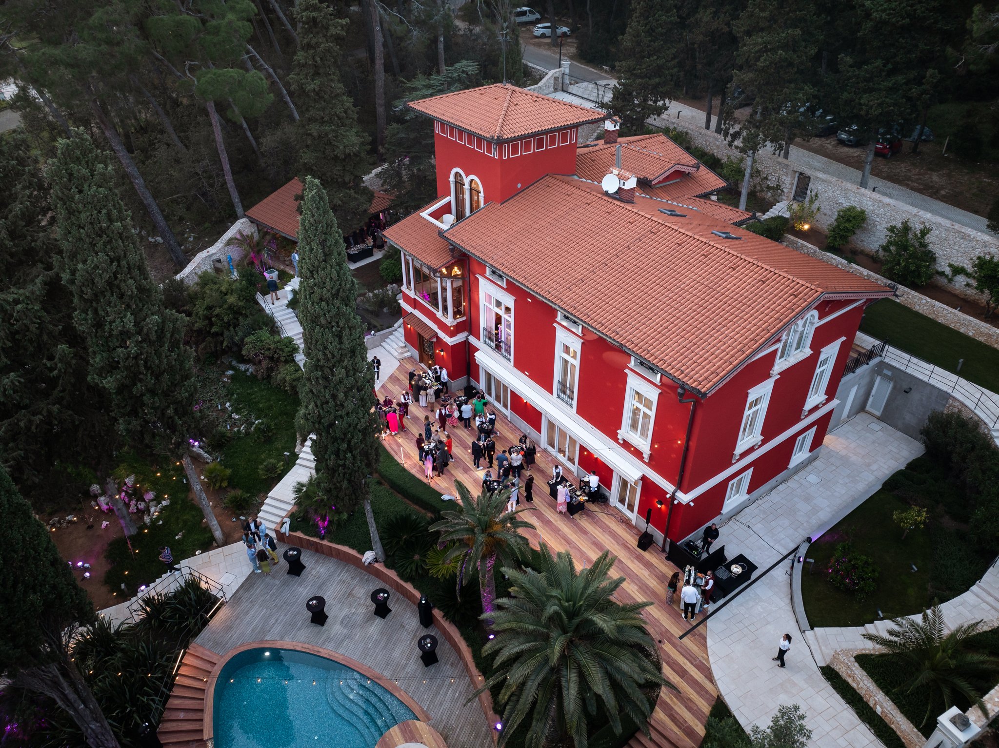 An outdoor gathering at a large, red house with tile roofs, surrounded by trees. There are guests mingling on the patio, near a swimming pool, with some standing around high tables, and a small band setup with musical equipment.