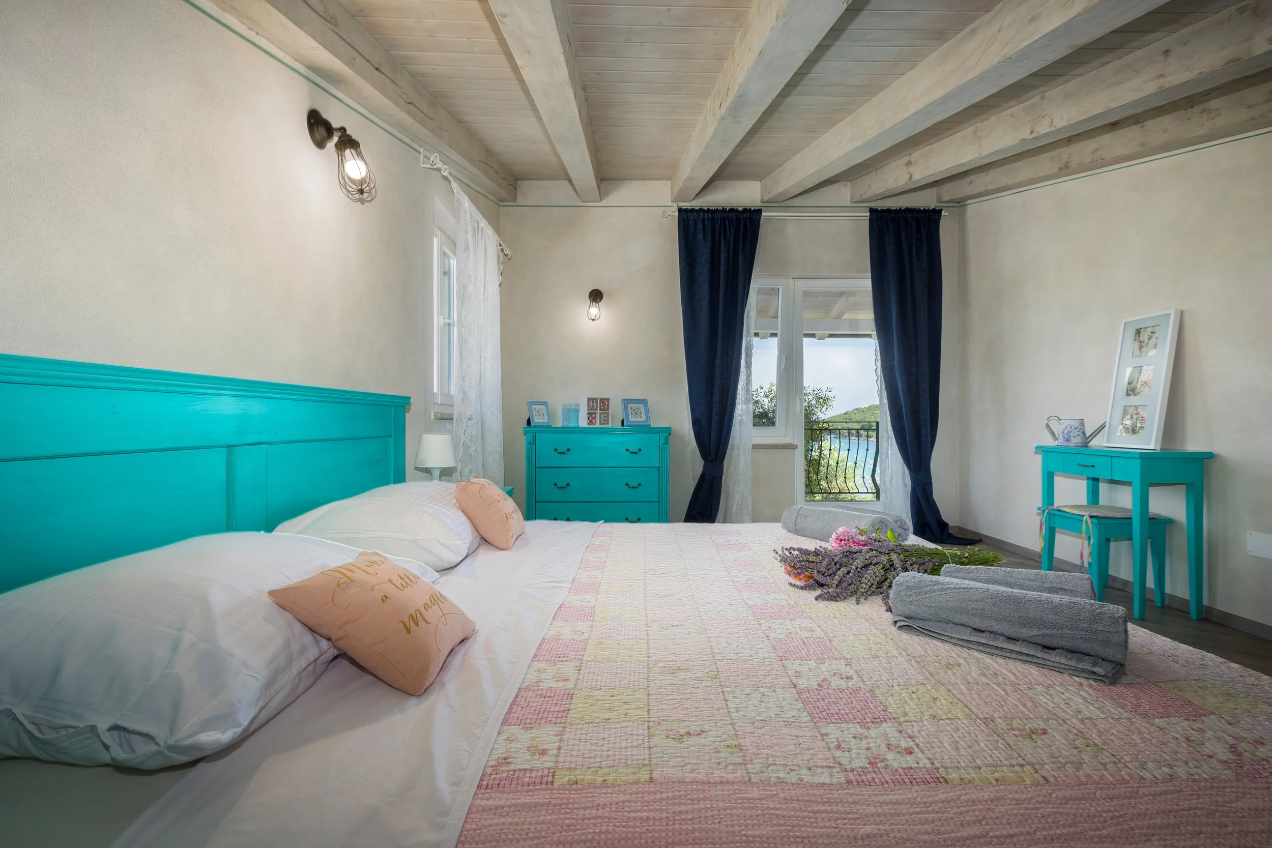 A cozy bedroom with a white and turquoise color scheme, featuring a bed with white pillows, a quilted bedspread, and decorative pillows with writing. The room has wooden beamed ceilings, a blue dresser, large windows with white and dark blue curtains