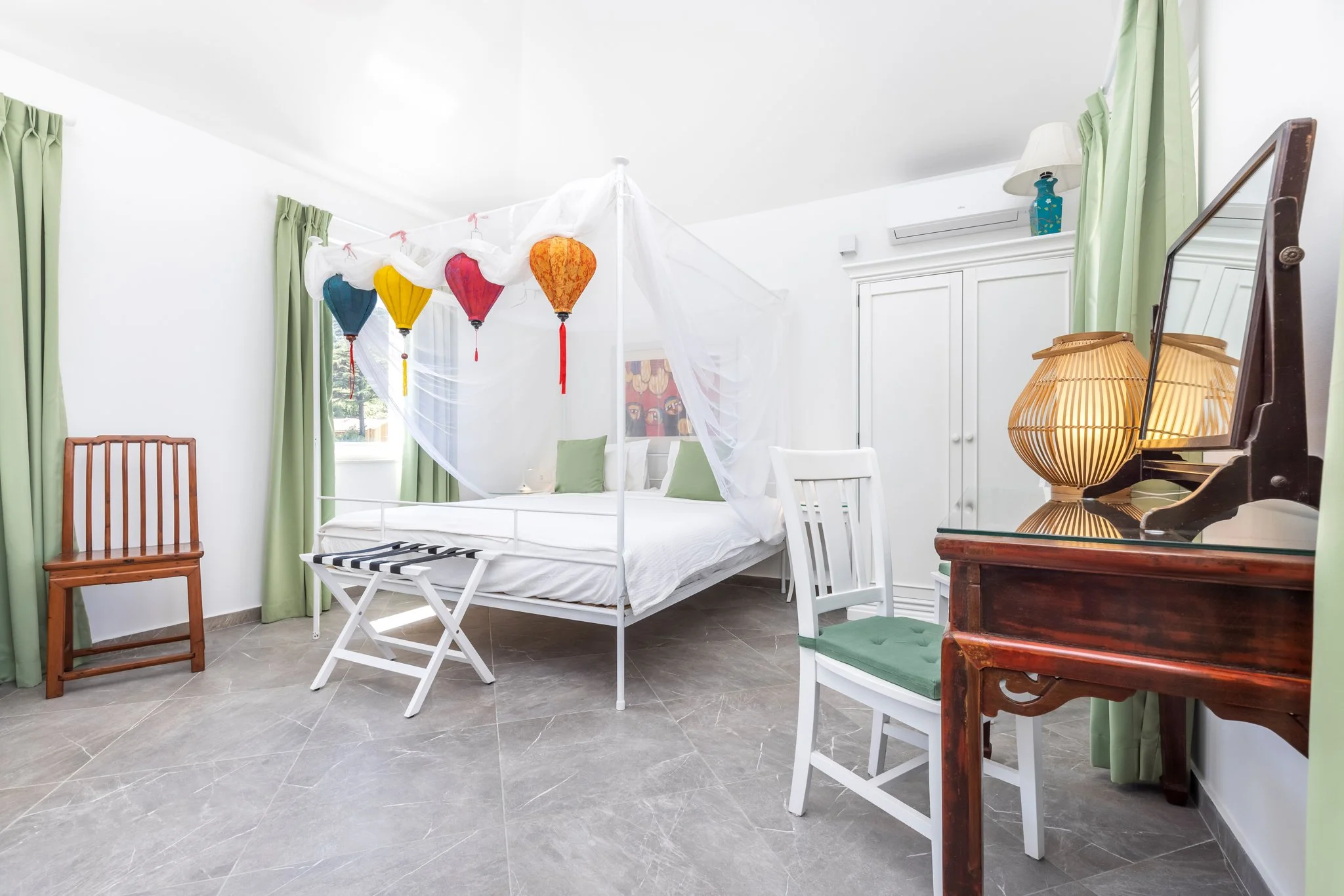 A bright bedroom with a white bed that has a canopy of sheer white fabric and colorful lanterns hanging from the ceiling. The room has white walls, light green curtains, and decorative furniture including a wooden chair, white chairs, a vintage dress