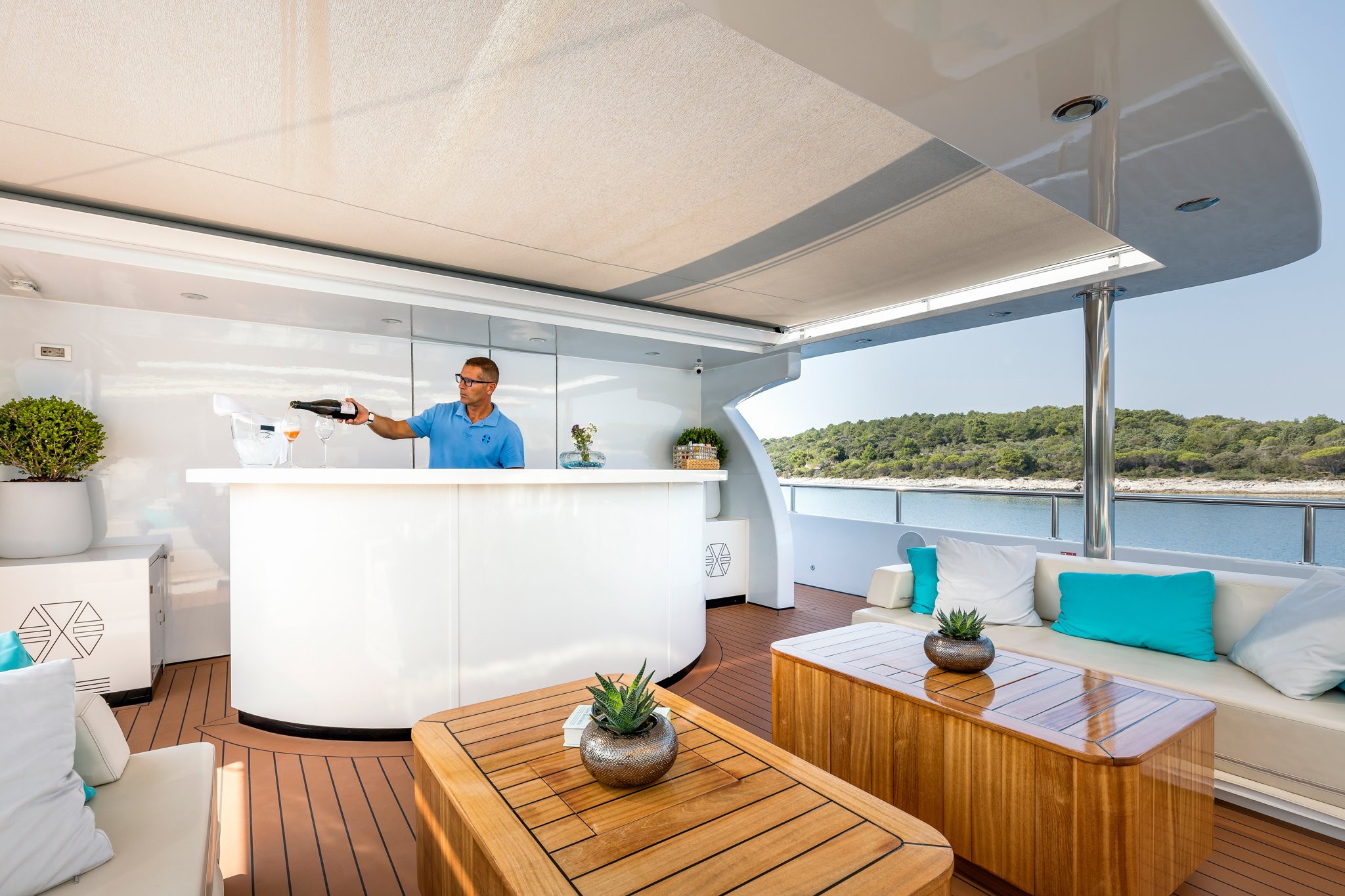 A man in a blue shirt serving drinks on a yacht with outdoor seating and greenery in the background.