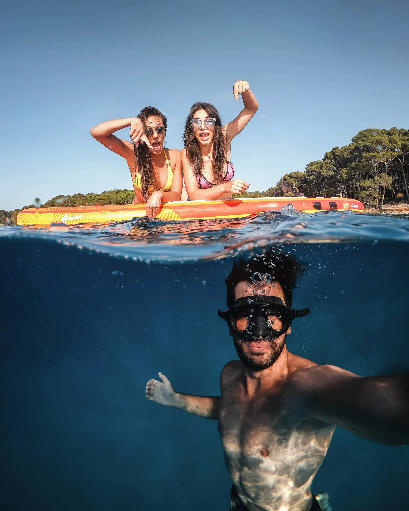 Two women sitting on an orange paddleboard in a body of water, making expressive gestures and wearing sunglasses, with a man in swimwear taking a selfie, and a forested shoreline in the background under a clear sky.