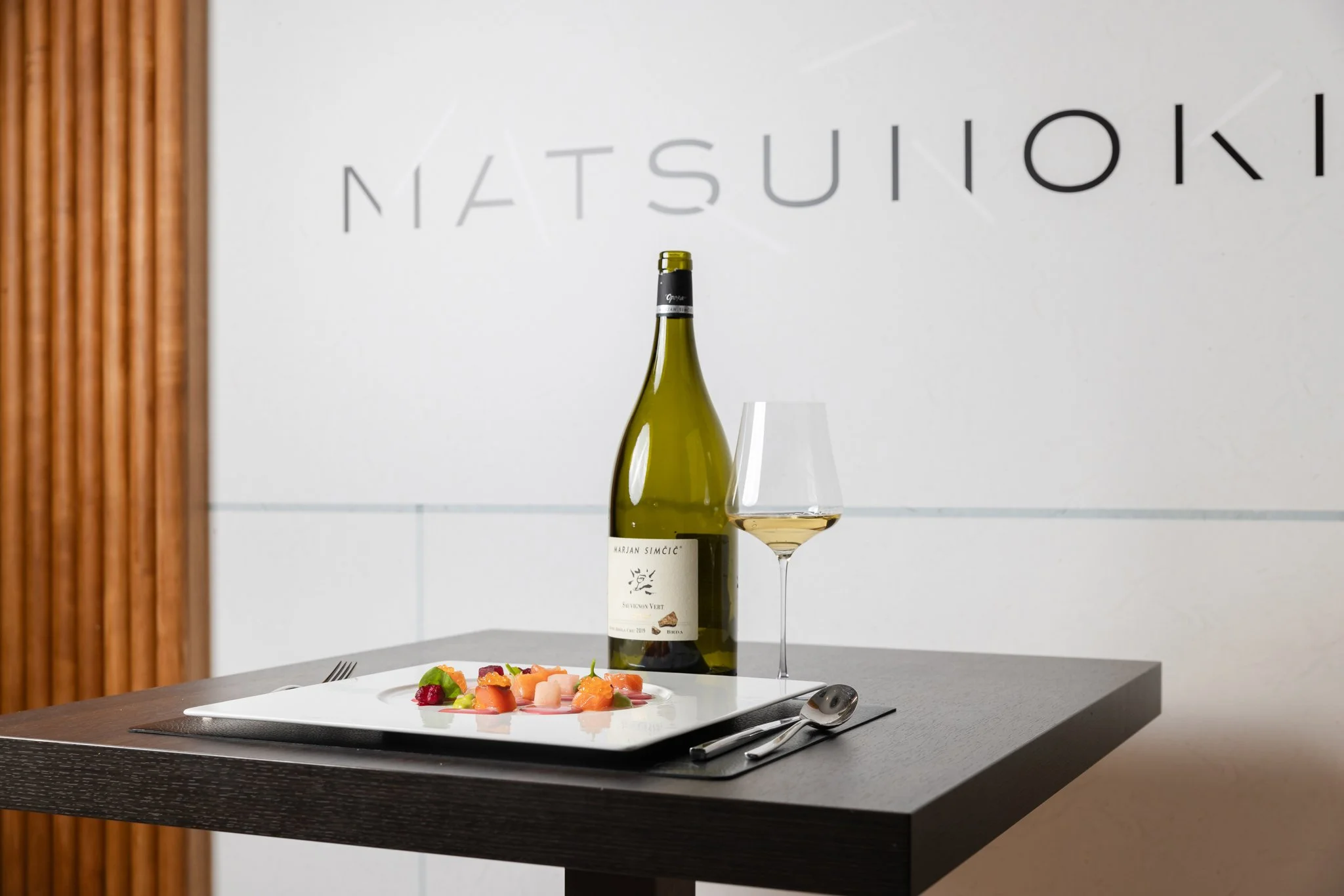 A wine bottle and a filled wine glass on a table with a plate of colorful food, such as salmon, vegetables, and garnishes, in a modern dining setting with a white wall and partial wooden paneling.