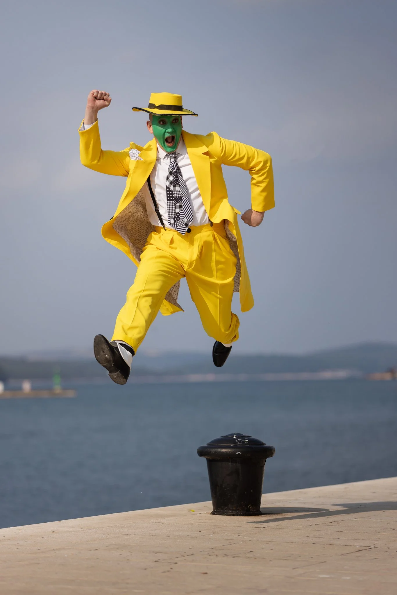 Person dressed as a clown with a yellow suit, striped tie, green face paint, and yellow hat jumping off a black bollard near water with a clear sky background.
