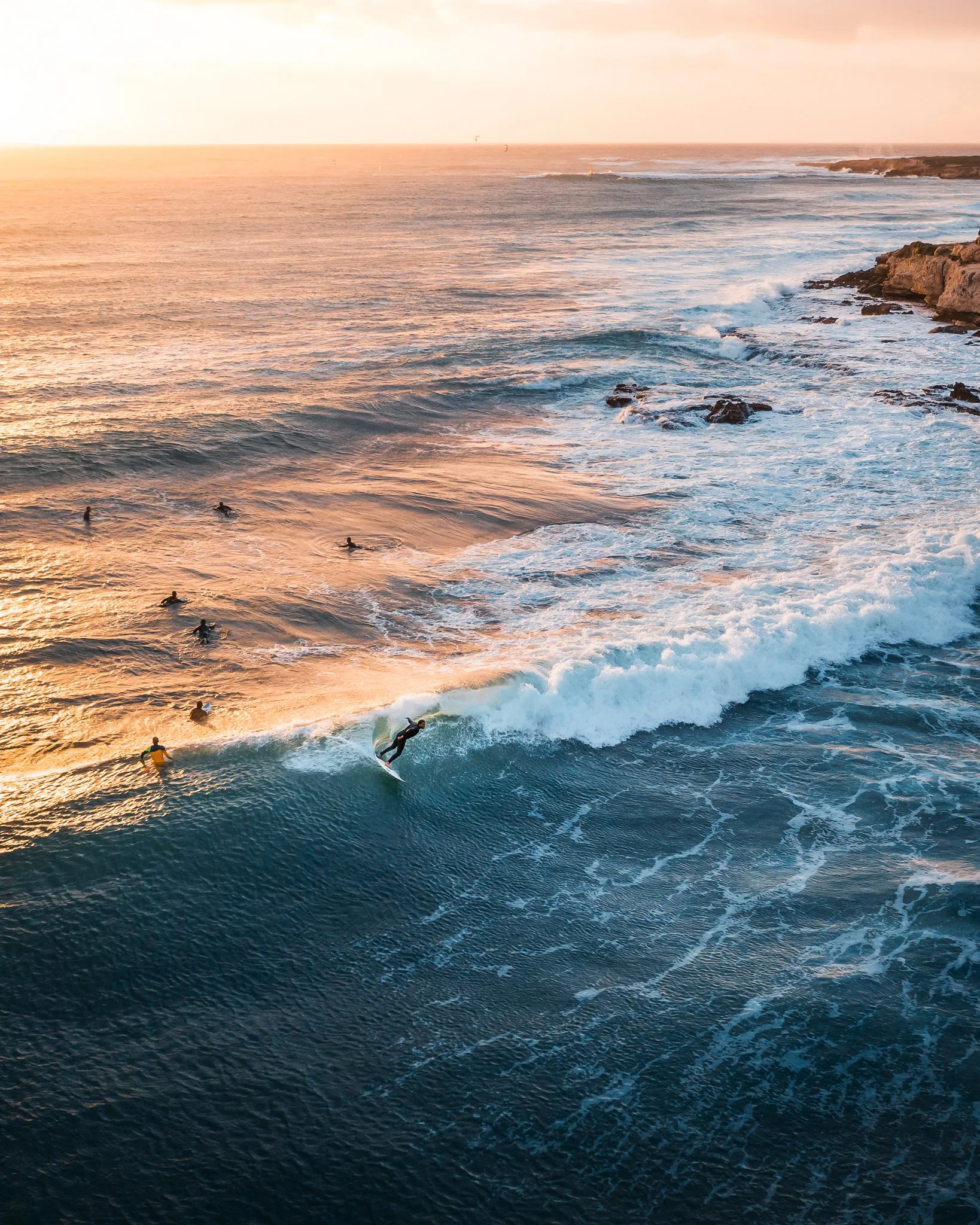 Sunset over the ocean with waves and a few surfers riding the waves near a rocky coastline.