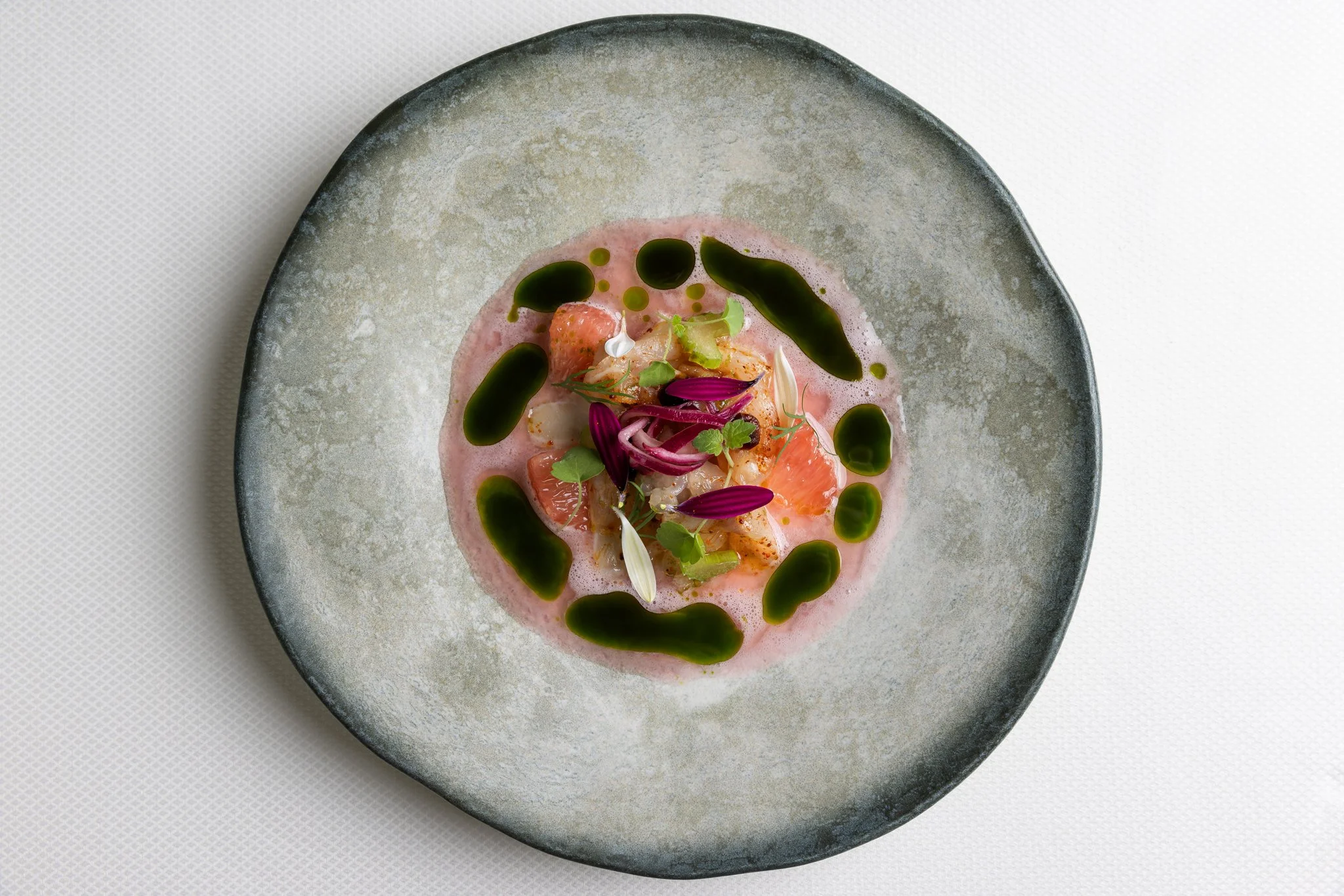 A gourmet dish with salmon, garnished with herbs, edible flowers, and served with a pink sauce and green oil droplets on a gray ceramic plate.