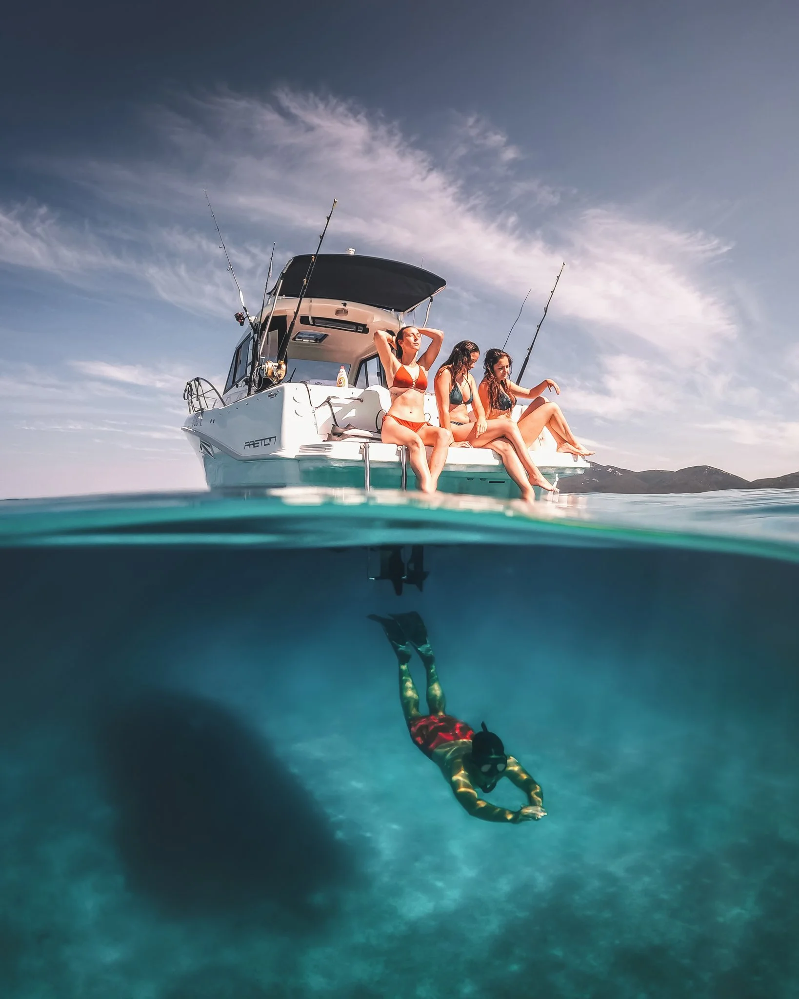 A boat floating on water with three women sitting on the edge, wearing swimsuits. One woman is sinking below the water, holding her hands together, while the other two women are sitting and relaxing on the boat.