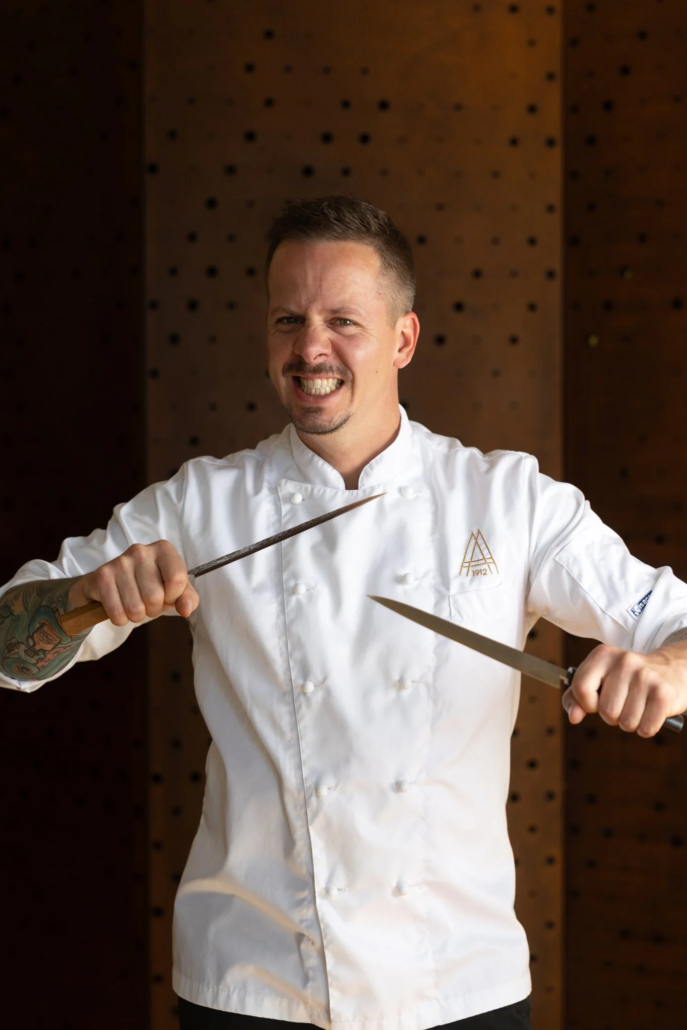 A smiling male chef with short brown hair and tattoos on his left arm, wearing a white chef's coat with a logo, holding a large kitchen knife in his right hand, in front of a brown wooden wall with small black pegs.