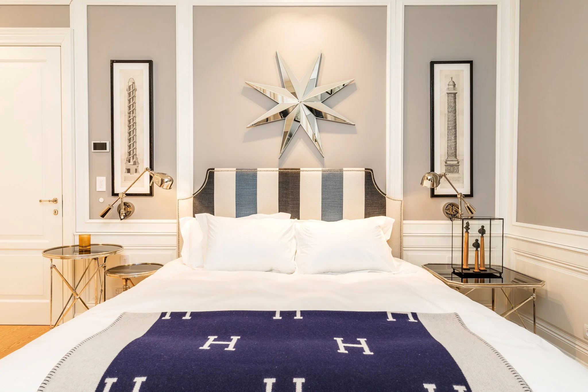A bedroom with a bed, a navy blue and gray 'H' rug, gold bedside lamps, framed art of towers on the wall, a large star-shaped mirror, and a black side table.