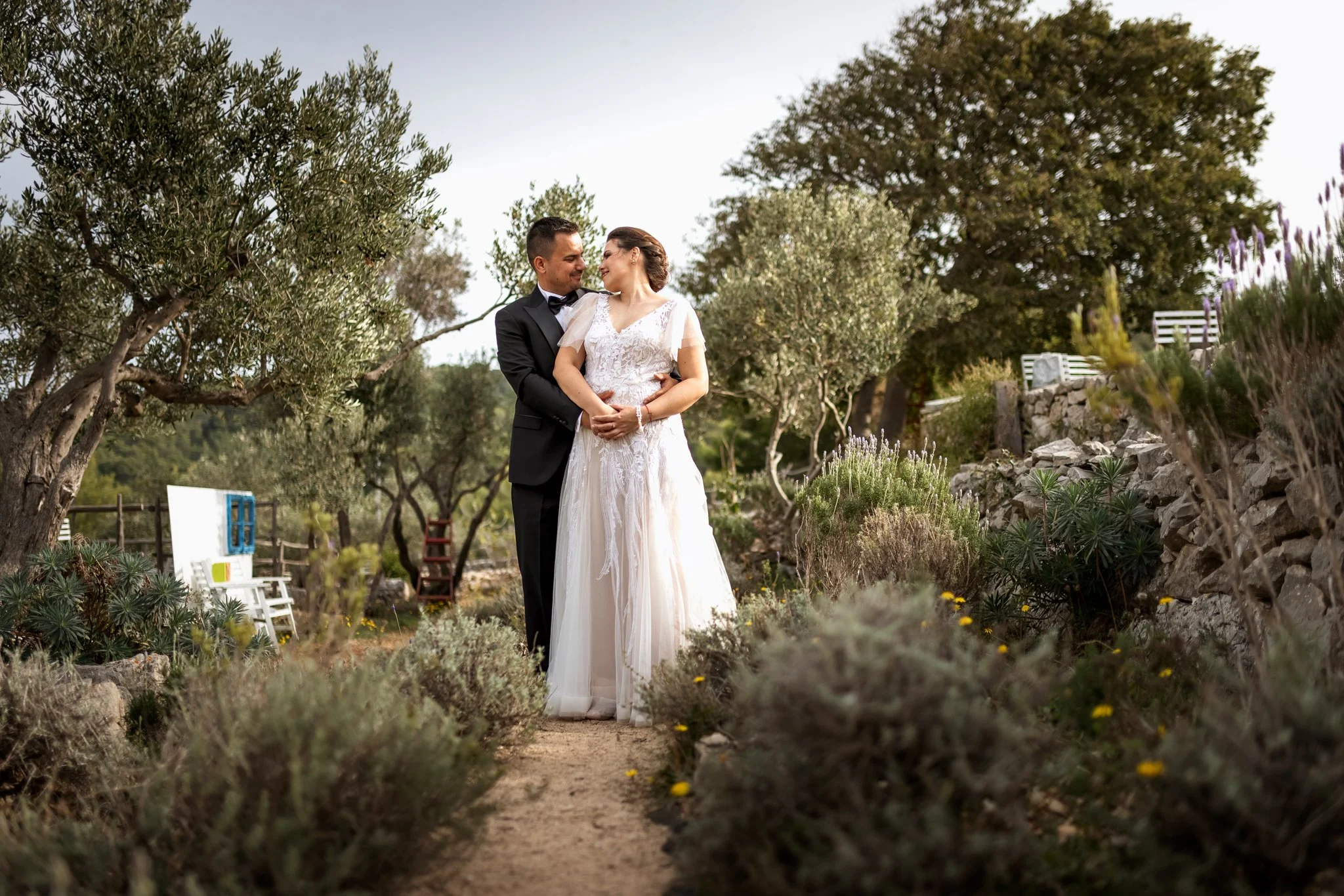 A bride and groom standing closely together outdoors in a garden setting with trees and plants, looking at each other tenderly.