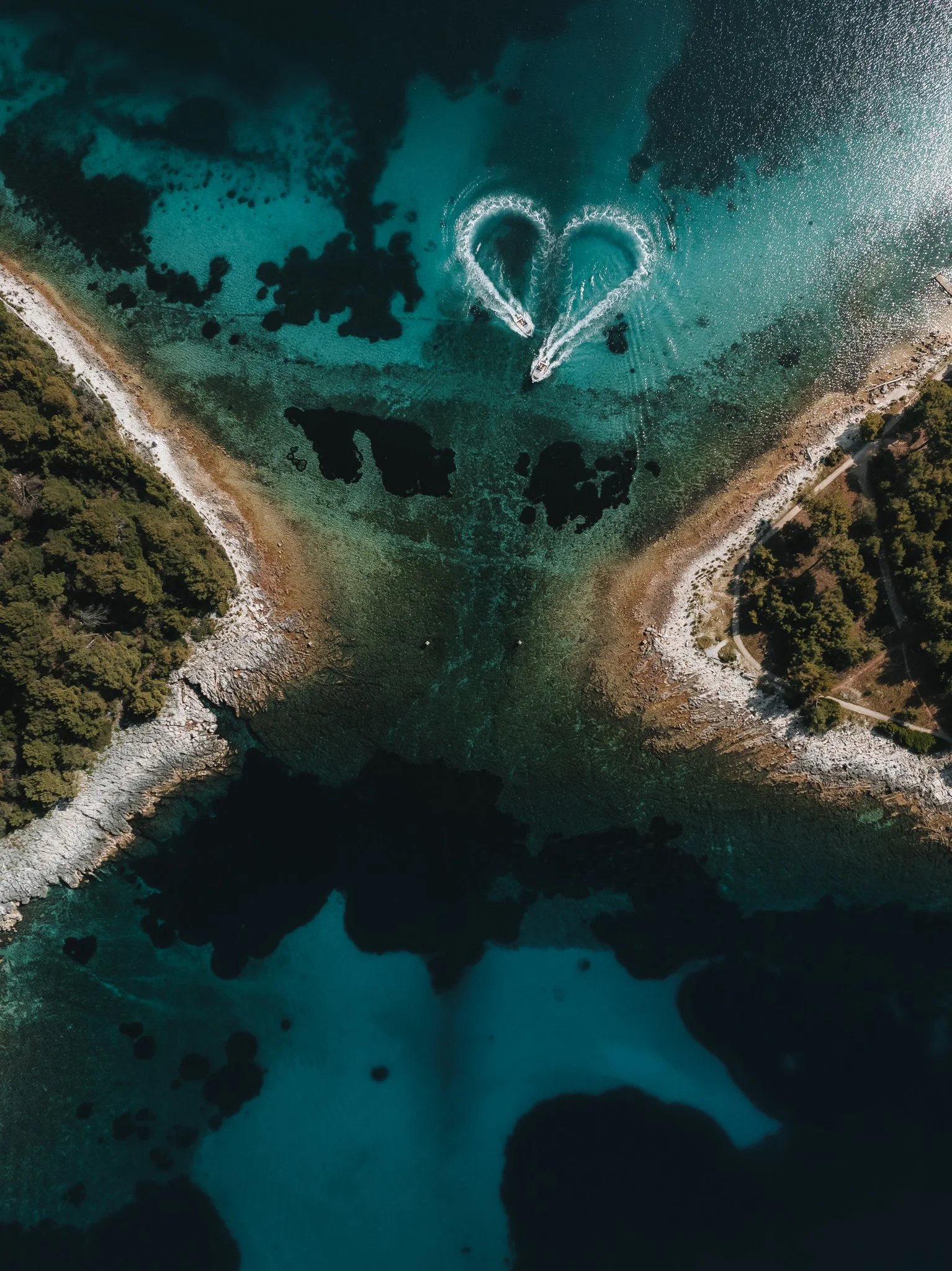 Aerial view of a coastal area with clear turquoise water, boats creating heart-shaped wakes, rocky shoreline, and trees along the coast.