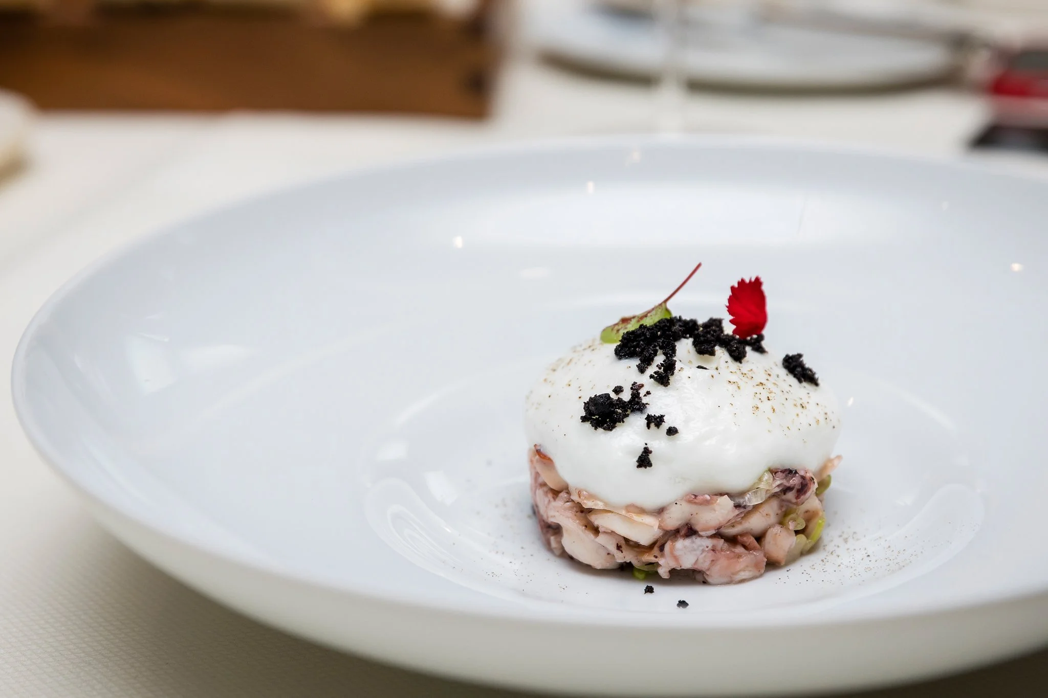 A plated dessert featuring a scoop of ice cream or mousse with a garnish of a red flower petal and black crumbs on top, served on a white plate.