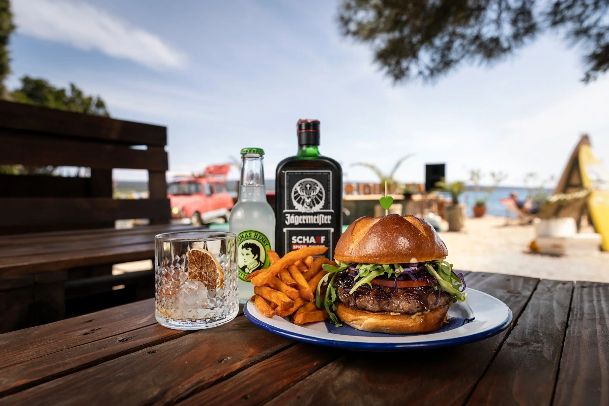 A plate with a burger, curly fries, and a glass of water with lemon slices on an outdoor wooden table.