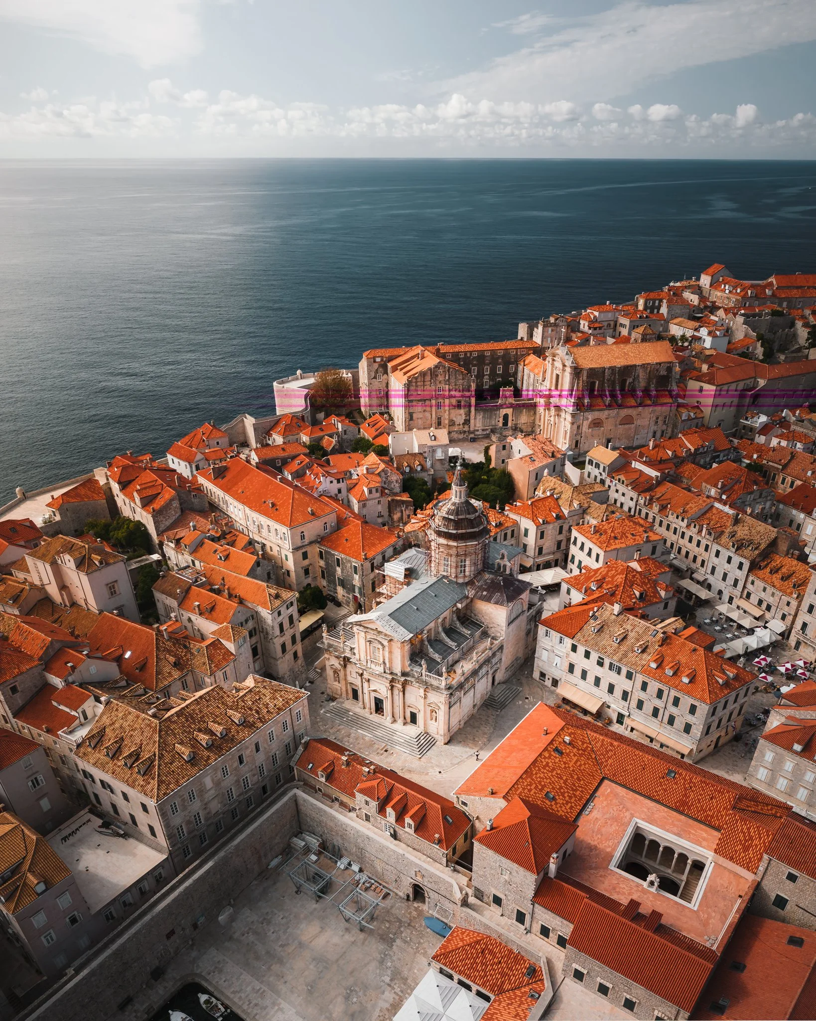 Aerial view of a historical European city with a large church or cathedral with a prominent dome, surrounded by orange rooftops and stone buildings, overlooking the sea with a partly cloudy sky.