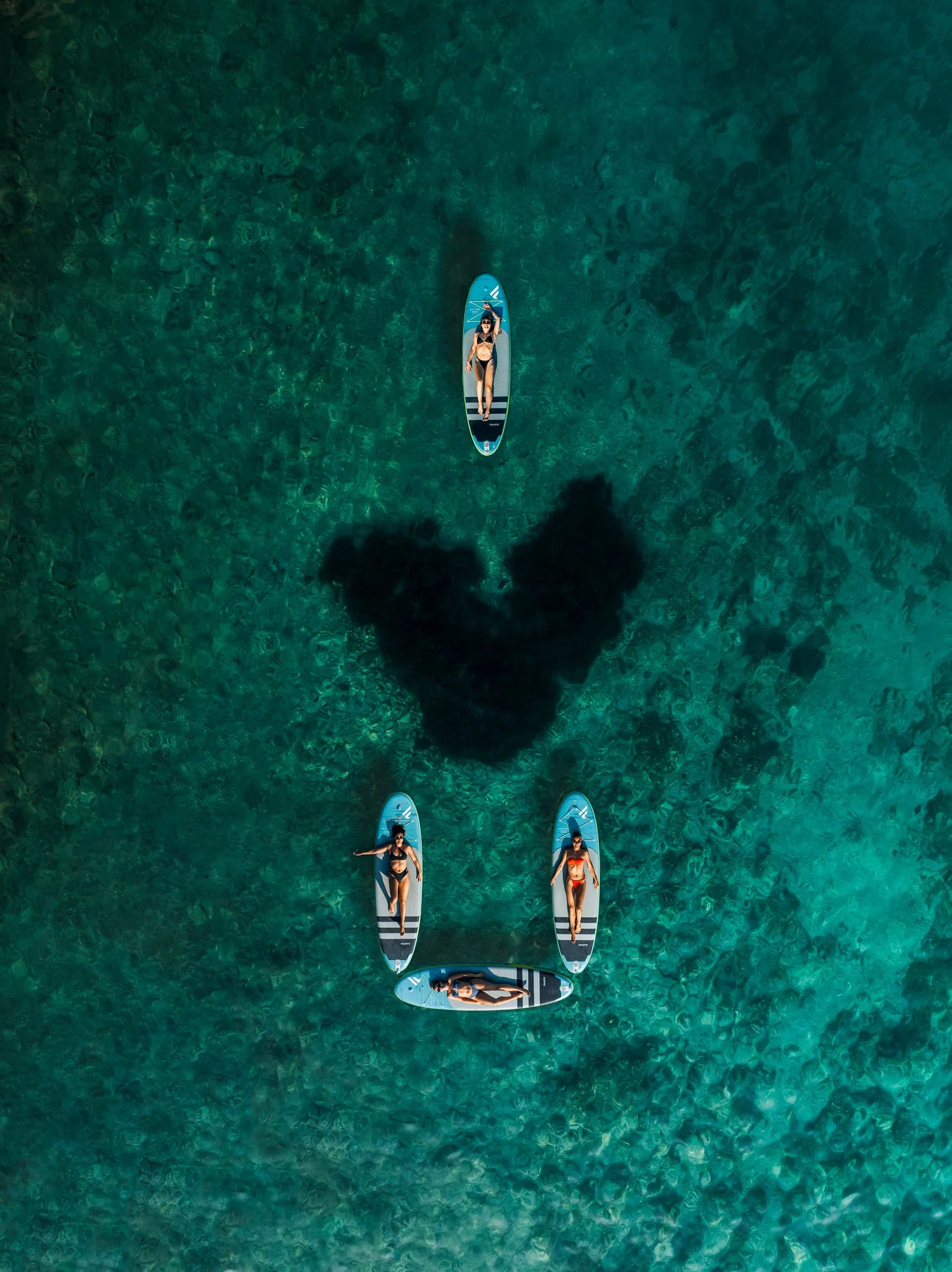 Four people are on paddleboards in clear, greenish water. Three women are standing on their boards, and one woman is lying down on her board. The formation of the boards and the sea grass looks like I love you.
