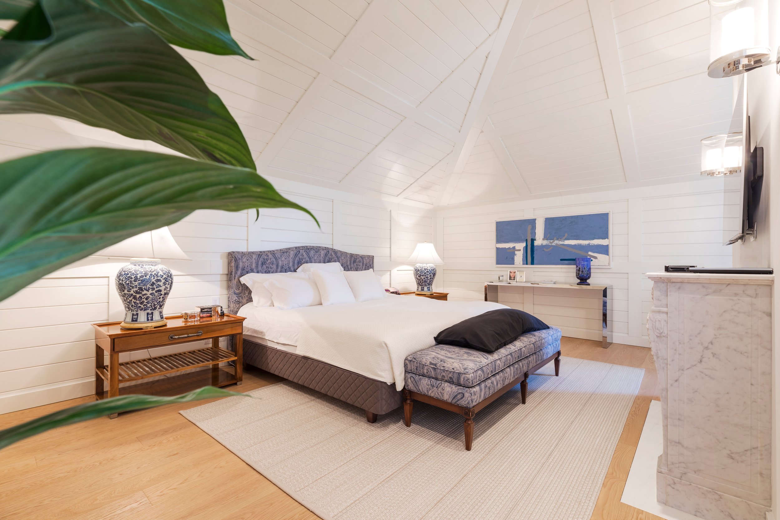 A cozy bedroom with white shiplap walls and ceiling, a large bed with a patterned upholstered headboard, white bedding, and two decorative blue and white lamps on wooden nightstands. There is a modern abstract painting on the wall, a small white desk