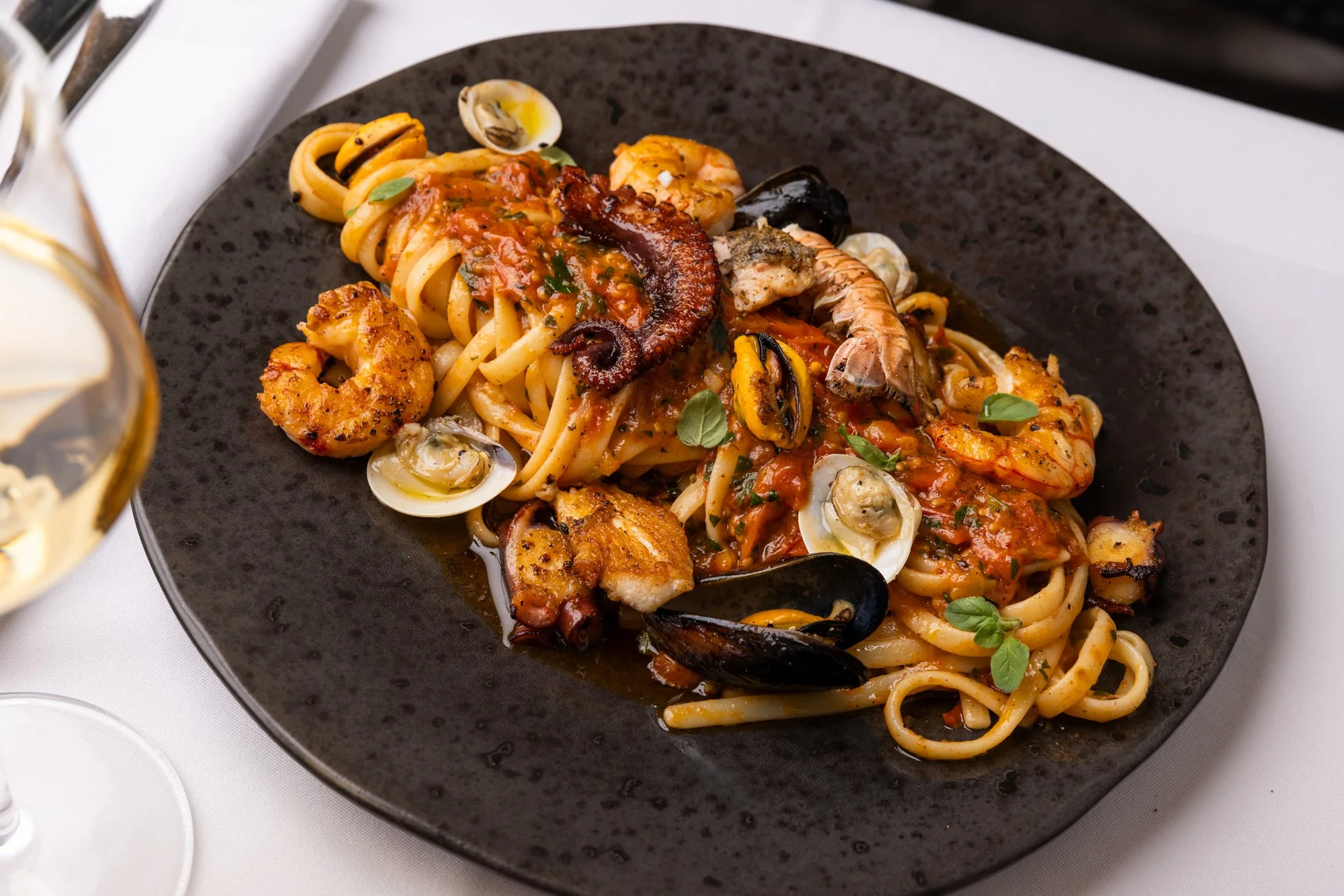 A black plate of seafood pasta with shrimp, mussels, and octopus in a tomato-based sauce, garnished with fresh herbs.