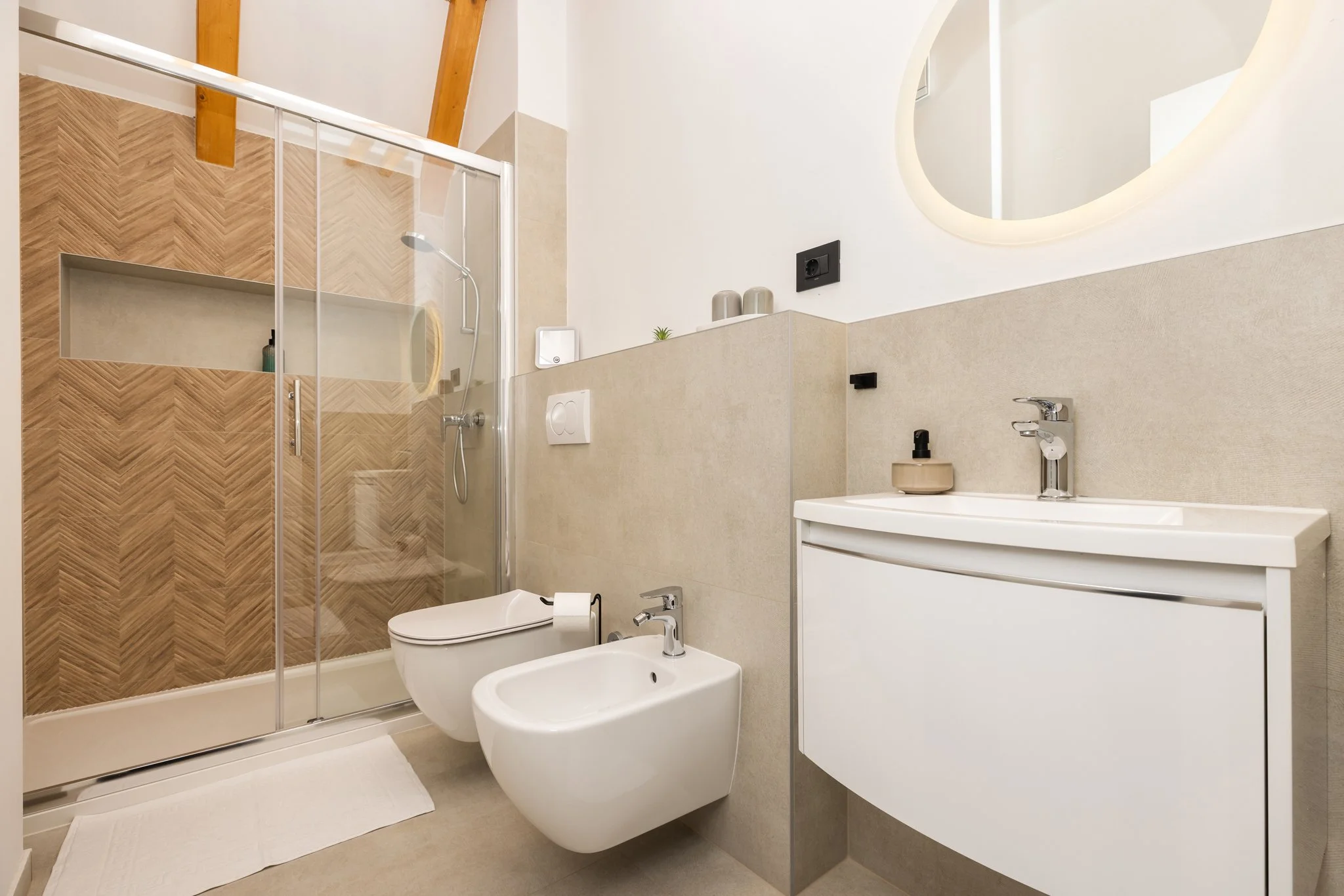 Modern bathroom with a glass-enclosed shower on the left, a wall-mounted toilet and bidet, a white sink with a curved mirror above, beige and wood tile accents, and minimal decor.