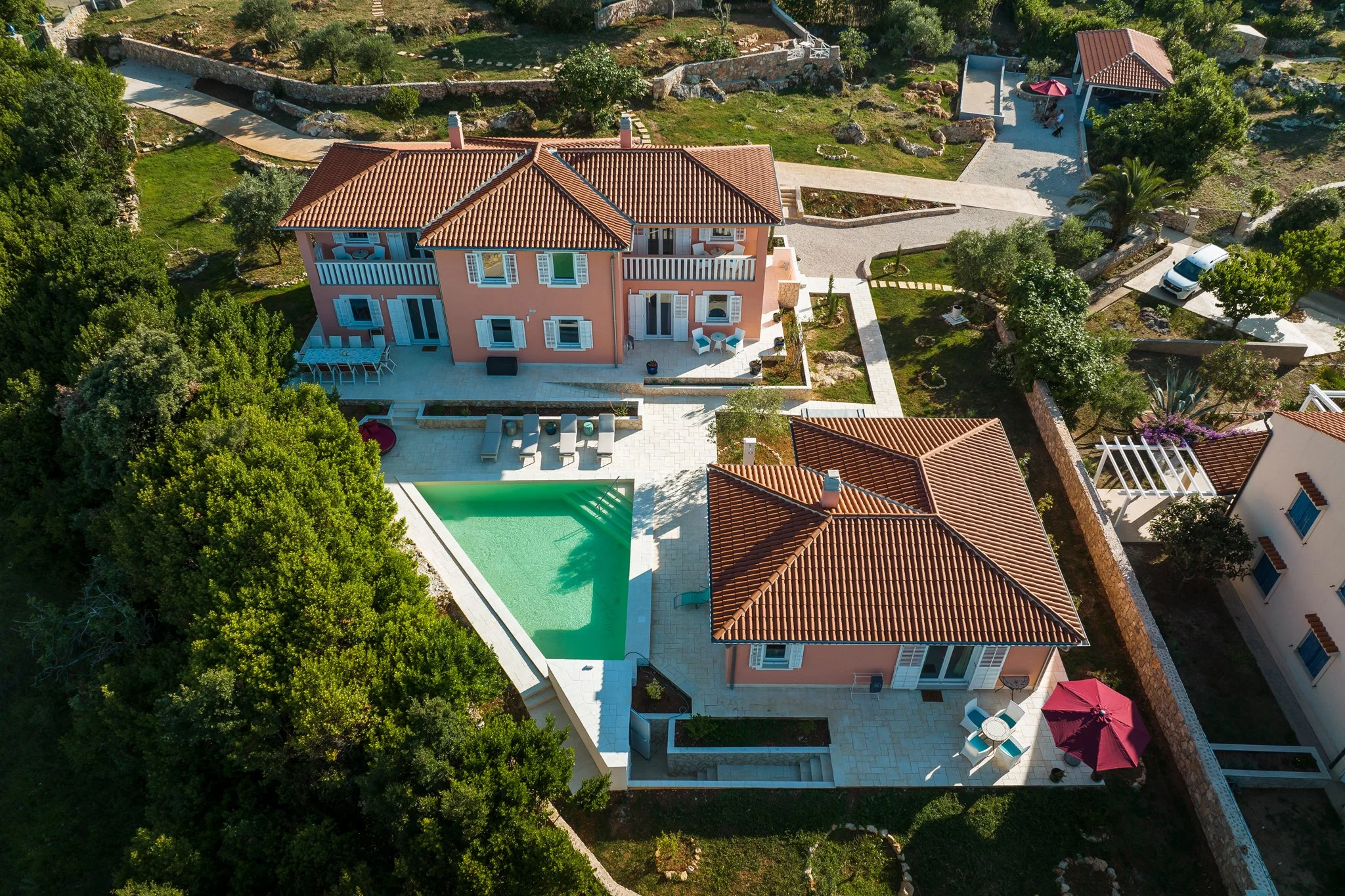 Aerial view of a pink house with a red-tiled roof, outdoor furniture, a swimming pool, and a well-landscaped yard with trees and pathways.