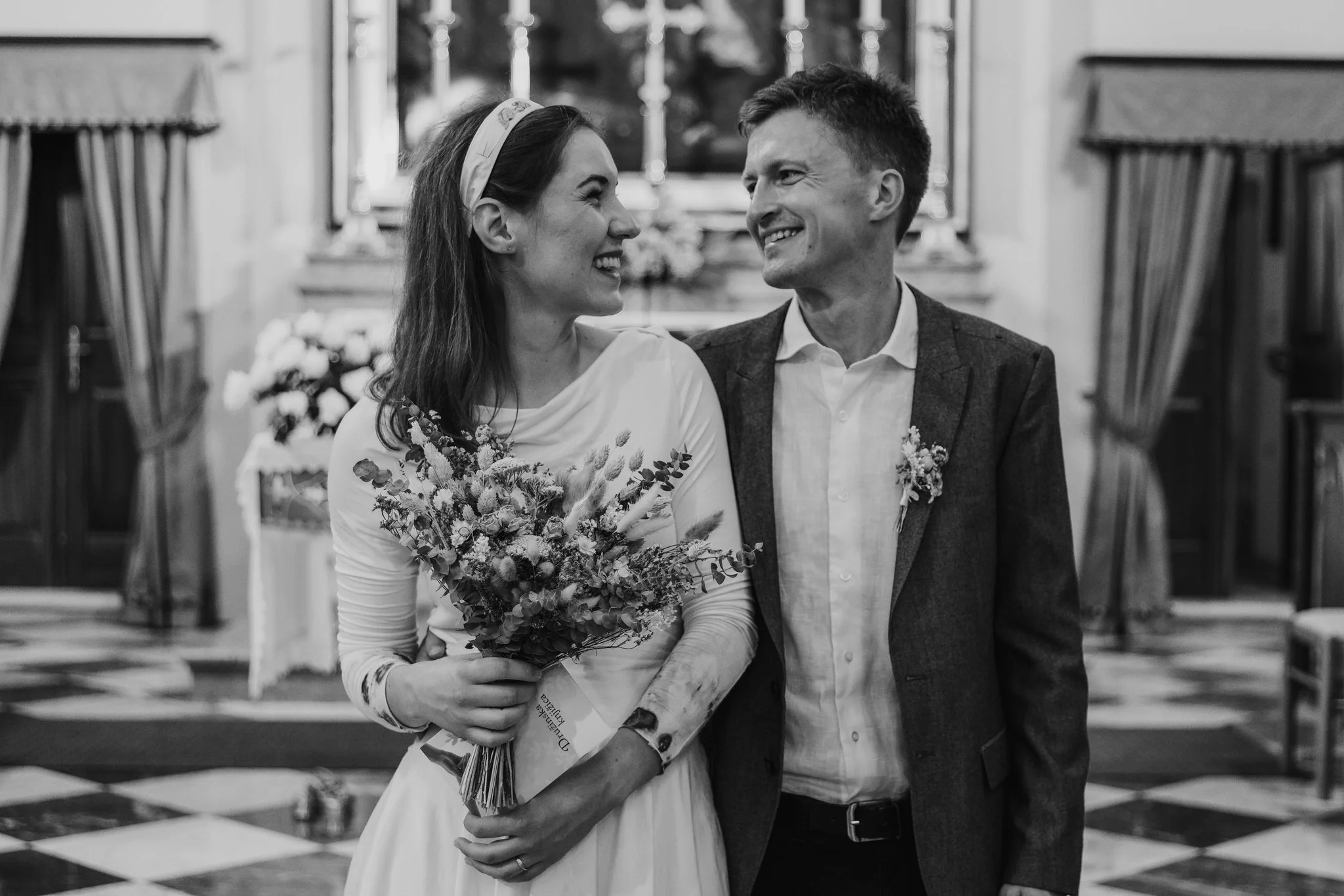 A smiling woman holding a bouquet stands next to a smiling man inside a church or chapel with altar and candles in the background.