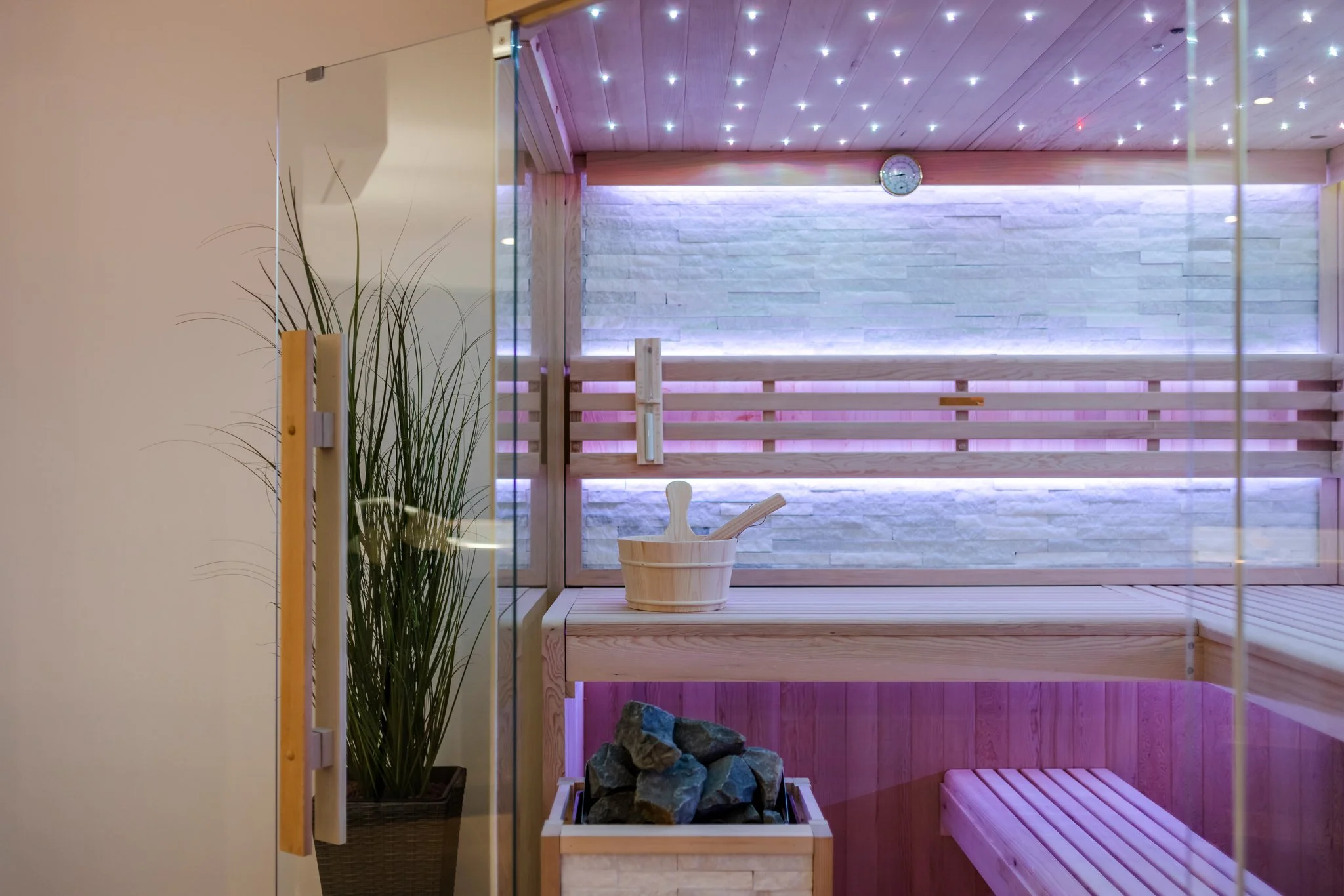 Interior of a sauna room with wooden benches, rocks, a bucket, and a wall with LED lighting.