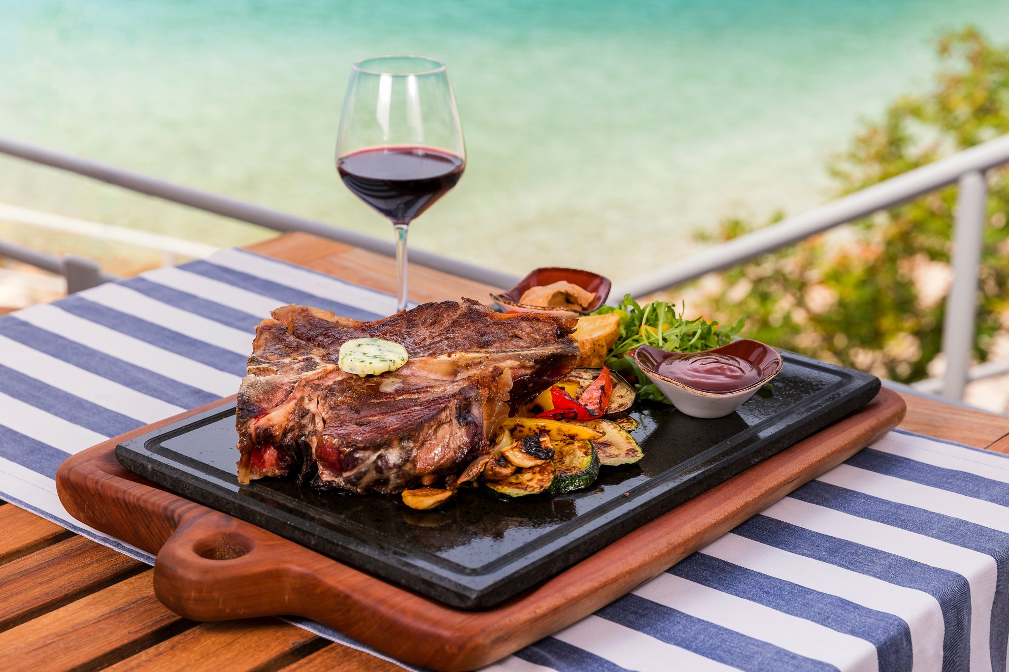 A grilled steak on a hot stone platter topped with herb butter, served with roasted vegetables and a side of sauces, accompanied by a glass of red wine, on a striped tablecloth overlooking water and greenery.
