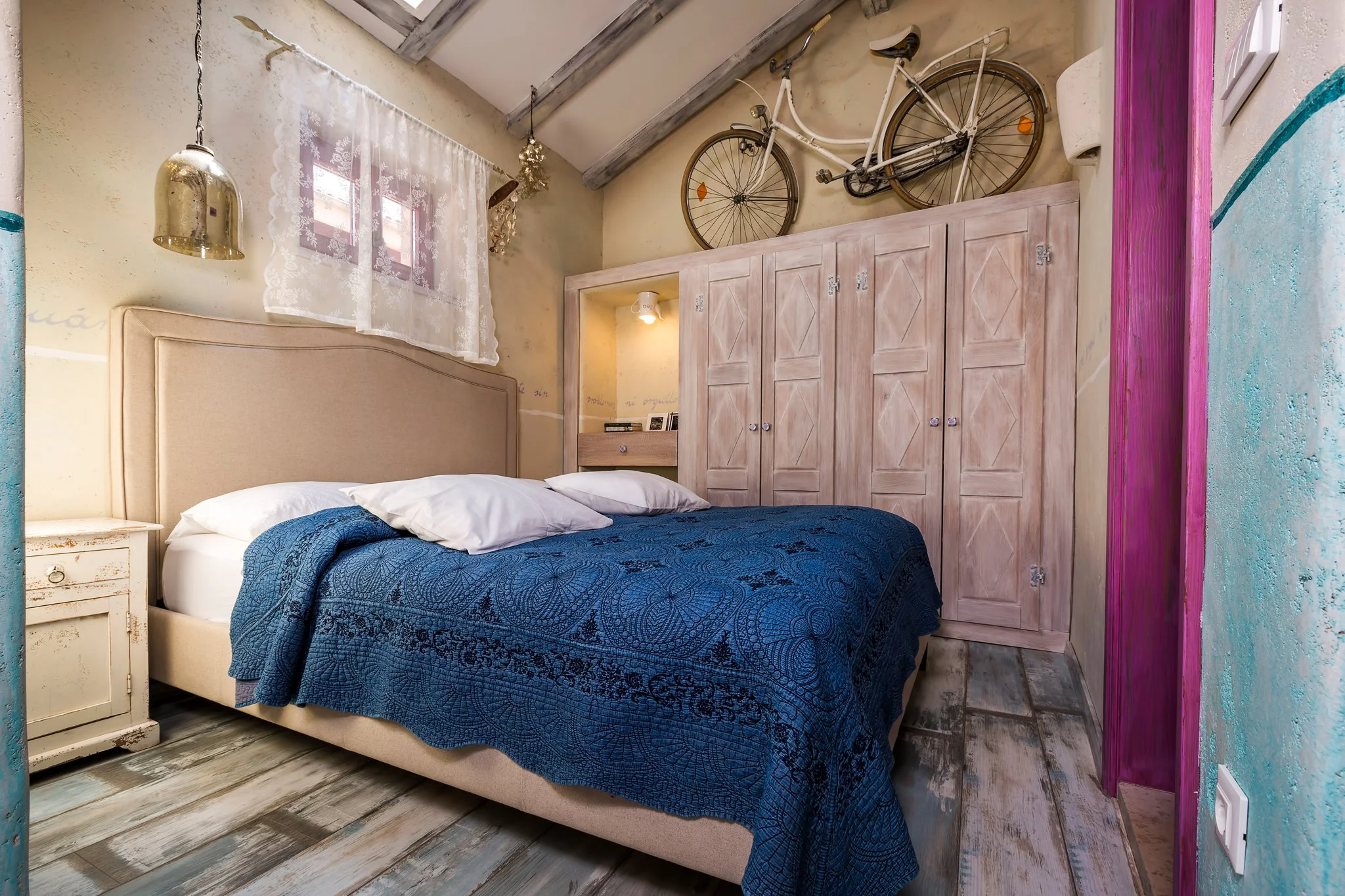 A cozy bedroom with a bed covered in a dark blue quilt and white pillows, a vintage nightstand, a wooden wardrobe with a bicycle mounted on the wall above it, and a small window with lace curtains. The room has a rustic and artistic decor with painte