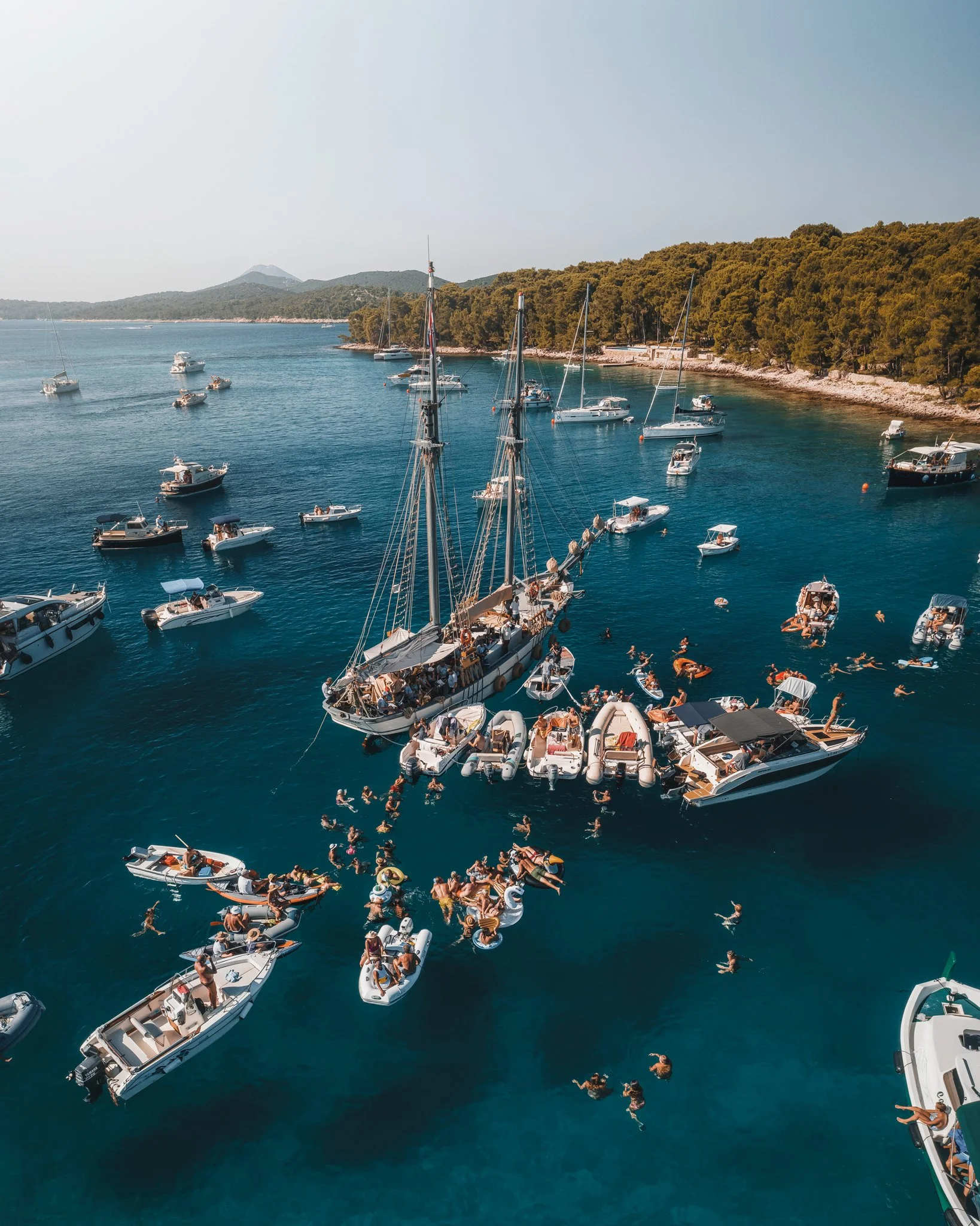 Aerial view of a crowded marina with boats and yachts, many people swimming and floating in the water, and a forested shoreline in the background.