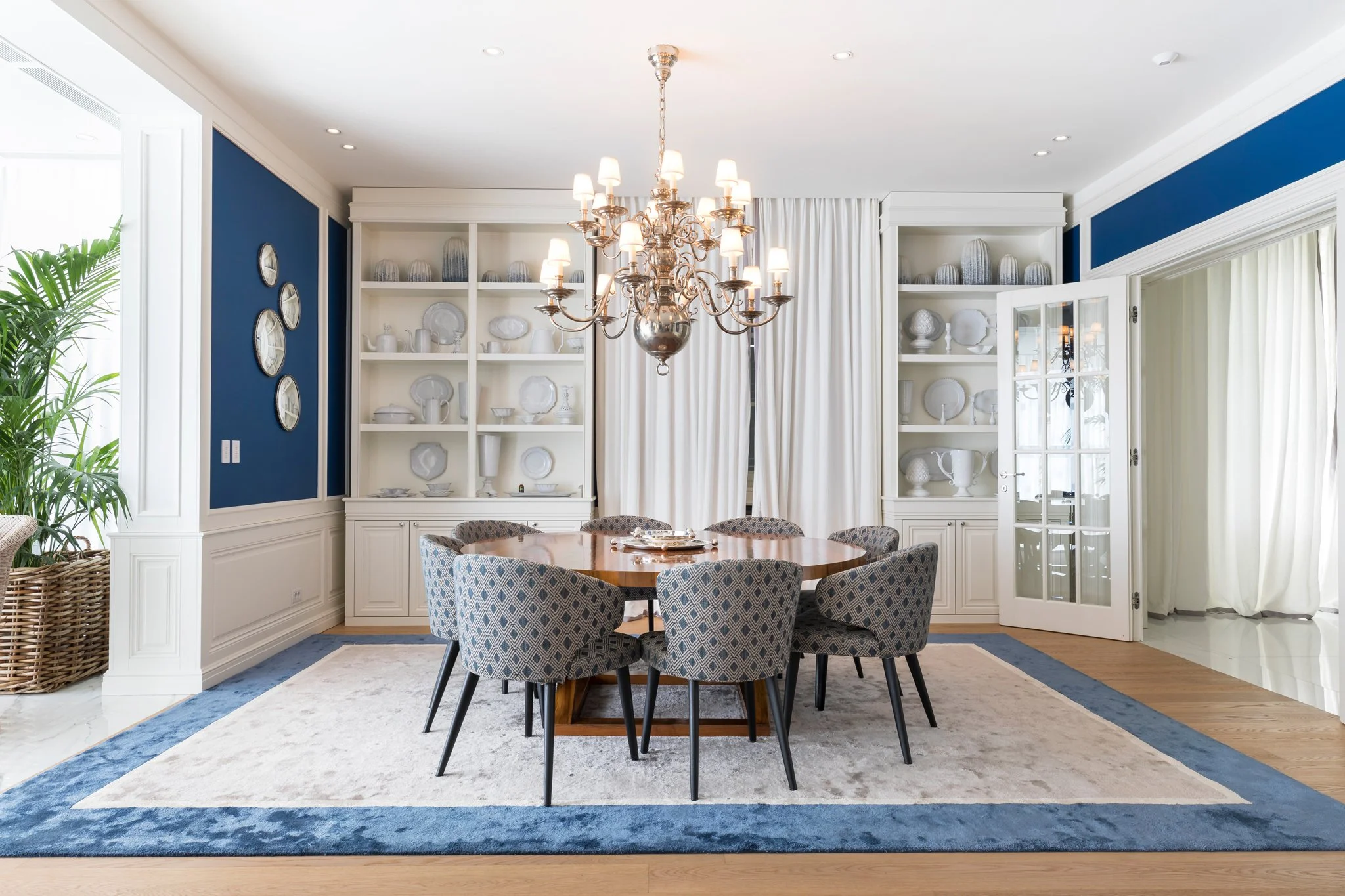 A dining room with a round wooden table surrounded by six patterned upholstered chairs. A large chandelier with multiple lit lamps hangs above the table. Behind, white built-in shelves display white dishware and decorative items. To the left, a tall 