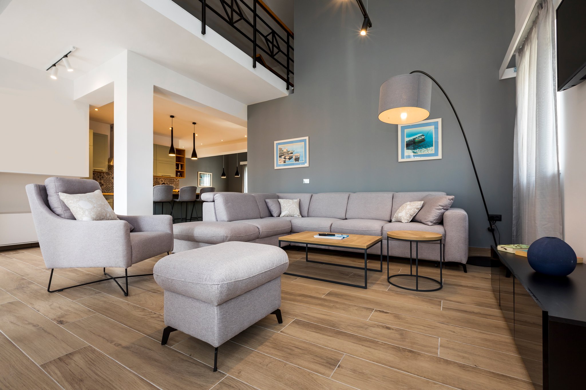 A modern living room with a light gray sofa, matching armchair, and a round ottoman, all on a wood-look tile floor. There are two small round tables near the sofa and a tall floor lamp with a gray shade. The blue wall behind the sofa has two framed p