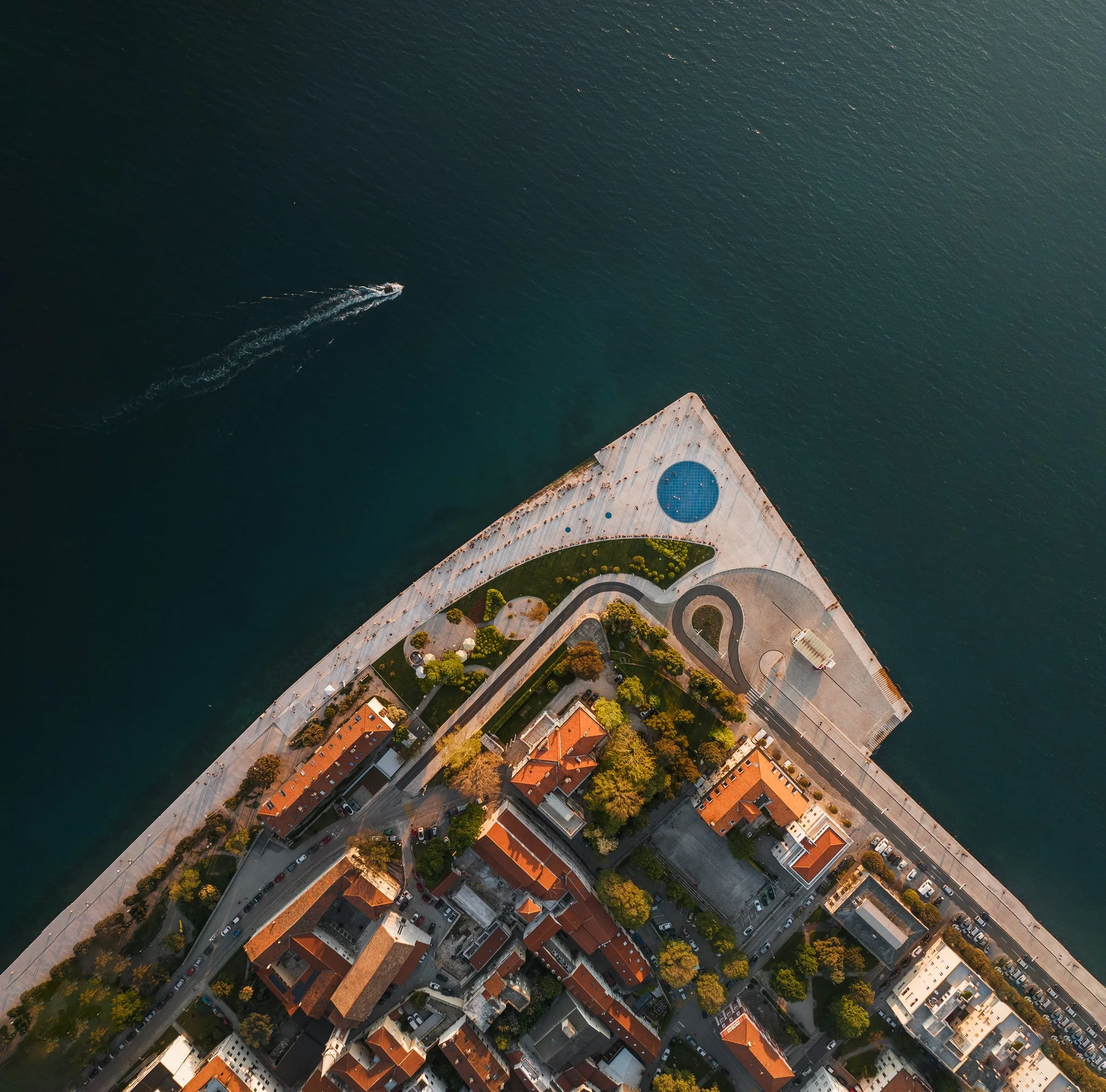A high-angle aerial view of a cityscape with residential houses, trees, roads, and parked cars along a large body of water. A boat is visible on the water, leaving a wake as it moves.
