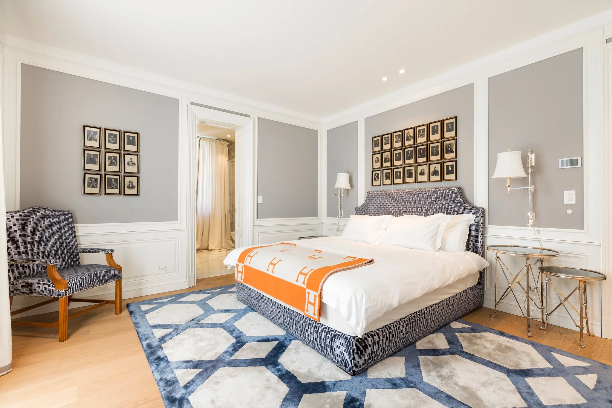 A bedroom with gray walls, framed black and white photos, a bed with a patterned headboard, white bedding, and an orange throw, two glass side tables, and a blue and white geometric area rug.