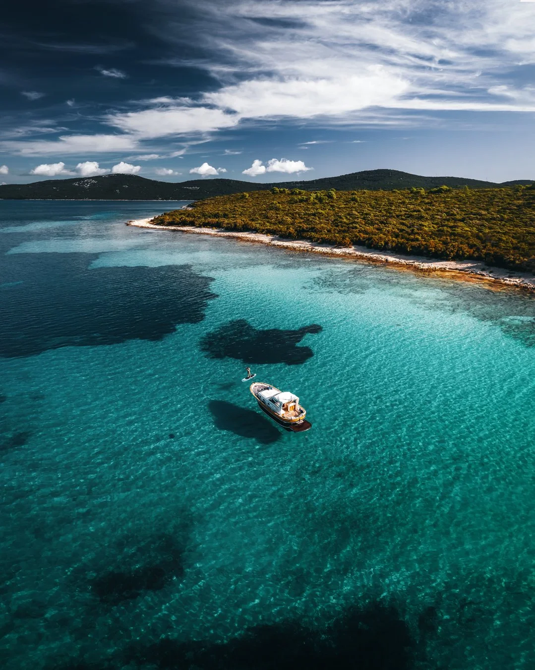 A boat floating on clear blue water near a coastline with green vegetation and hills in the background under a partly cloudy sky.
