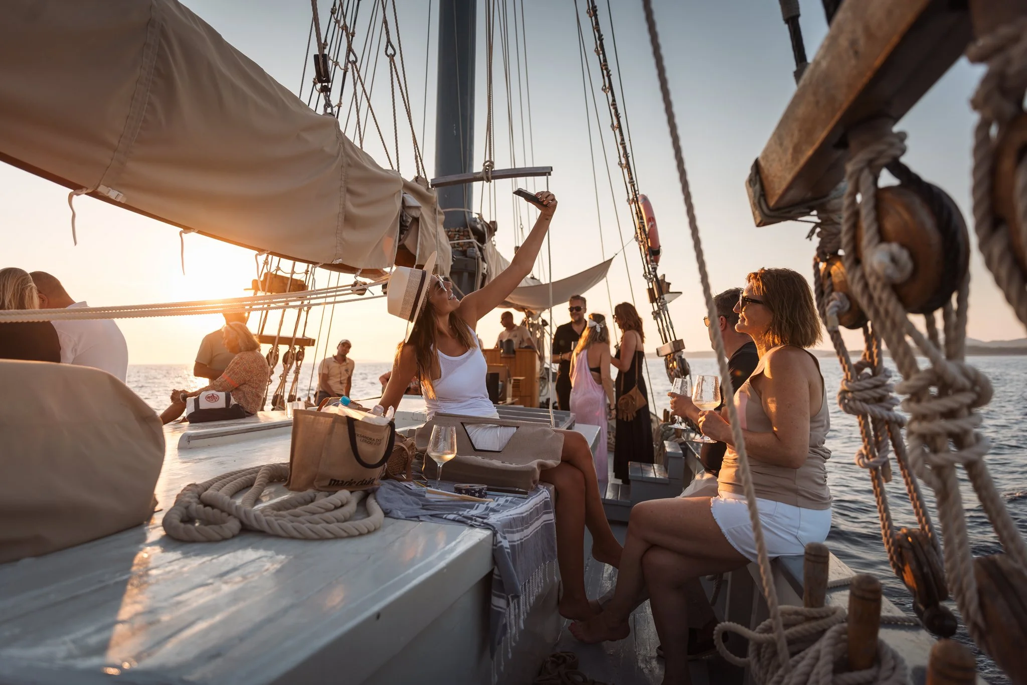 People enjoying a sunset on a sailboat with wine glasses. Some are sitting while others stand, taking photos. The boat has ropes and a sail, with a calm sea in the background.