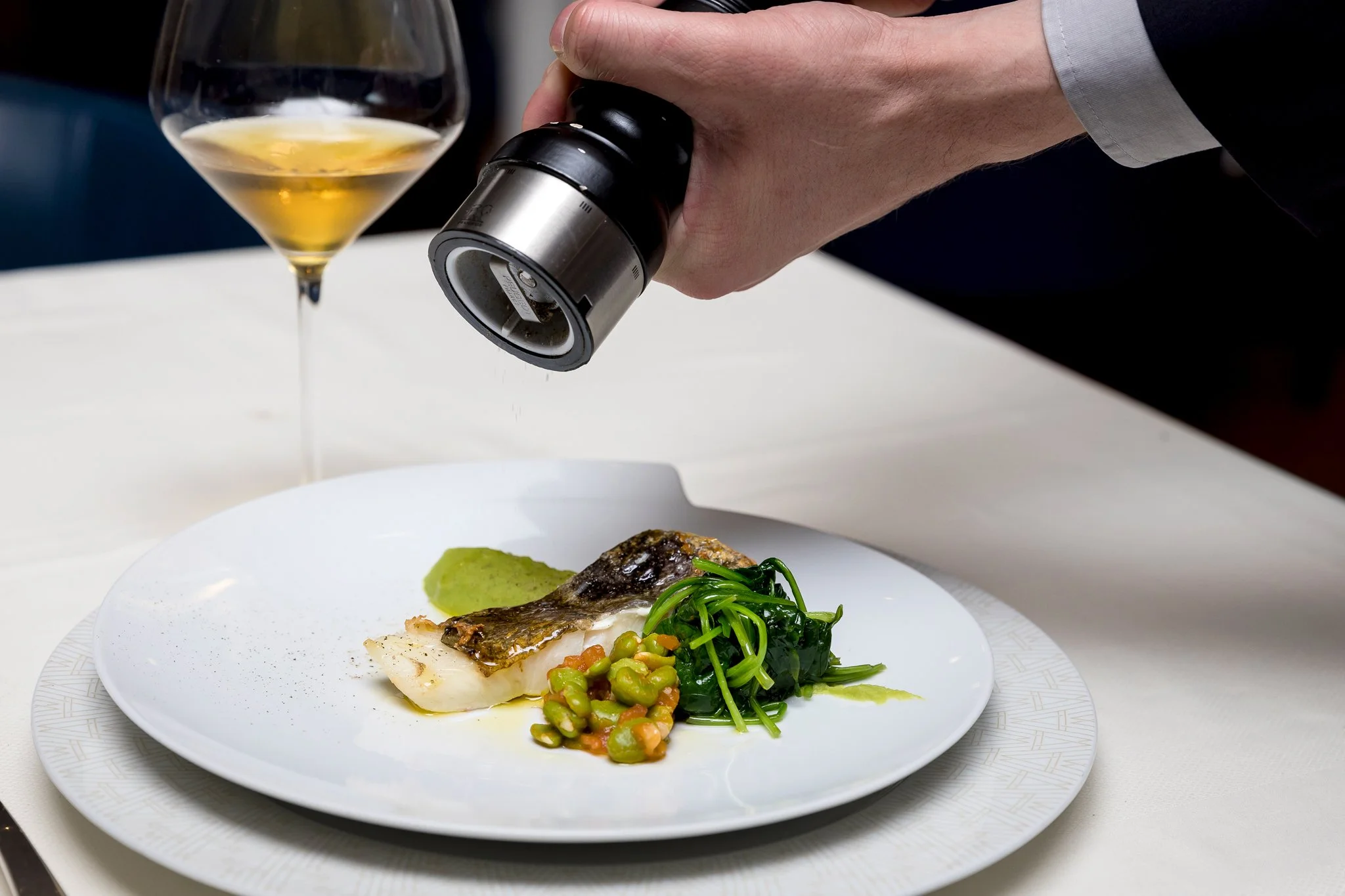 Chef seasoning plated fish dish with greens and vegetables, wine glass in background.