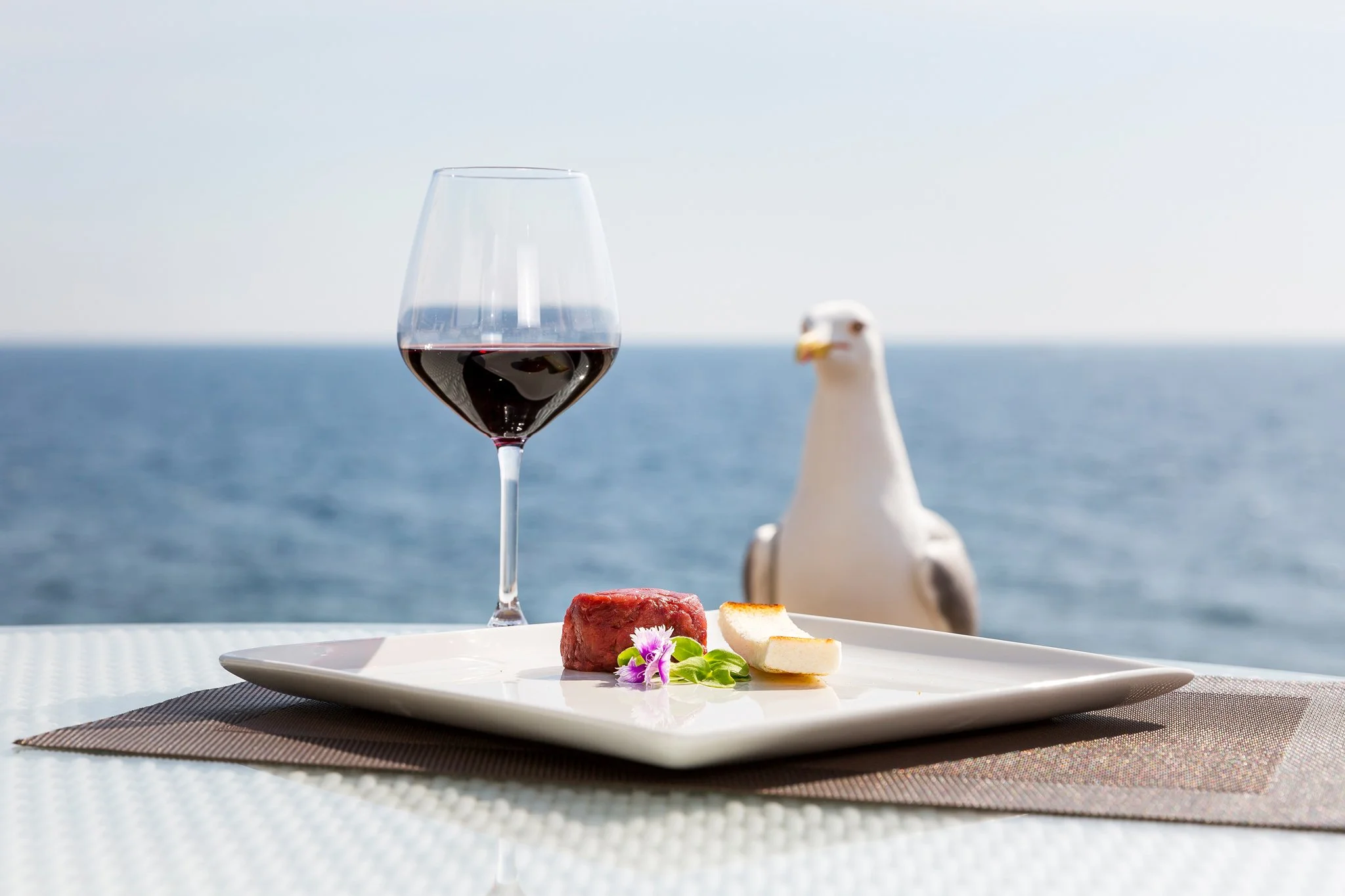 A glass of red wine on a table with a seaside view, alongside a plate of cheese, a piece of meat, and some decorative flowers, with a seagull in the background.